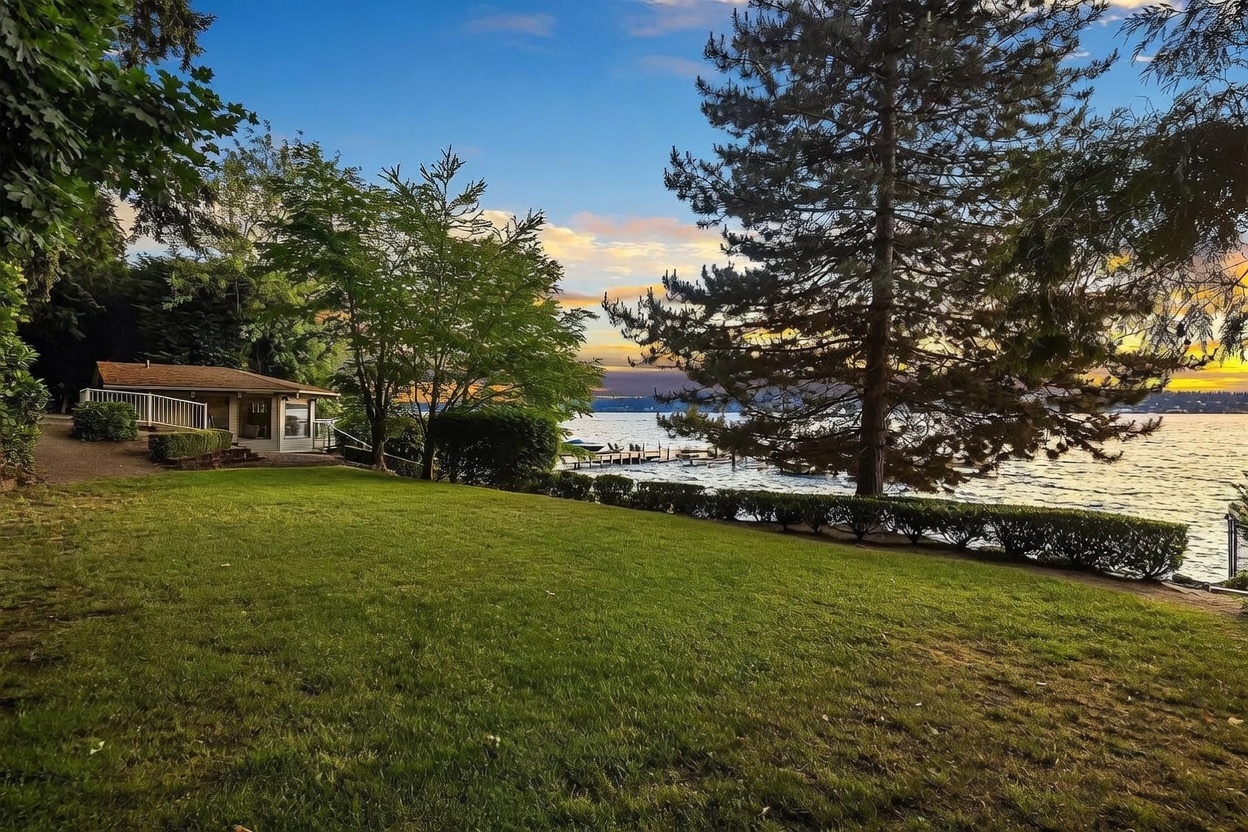 This scenic backyard view captures a lush, expansive green lawn leading toward a tranquil waterfront setting at sunset. A modest, single-story home with a deck sits to the left, framed by mature trees and a manicured hedge that borders the water's edge. The perspective is wide and inviting, showcasing the peaceful connection between the private property and the lake beyond.
