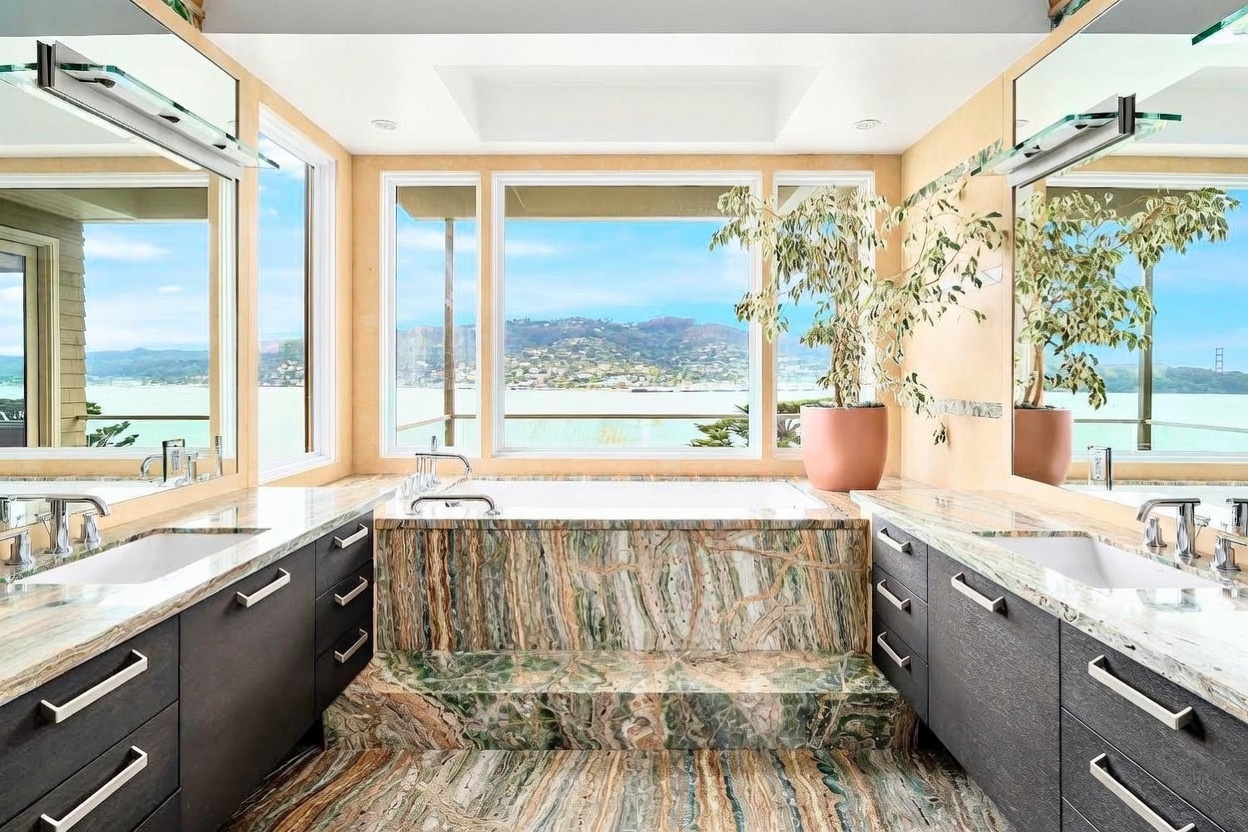 This luxurious primary bathroom features a striking, multi-toned stone tub surround and matching flooring that creates a dramatic focal point. The space is symmetrically designed with dual dark-toned vanities on either side, each topped with light-colored stone counters and large mirrors. A large window behind the soaking tub provides an expansive, panoramic view of the water and distant hills, filling the room with natural light.