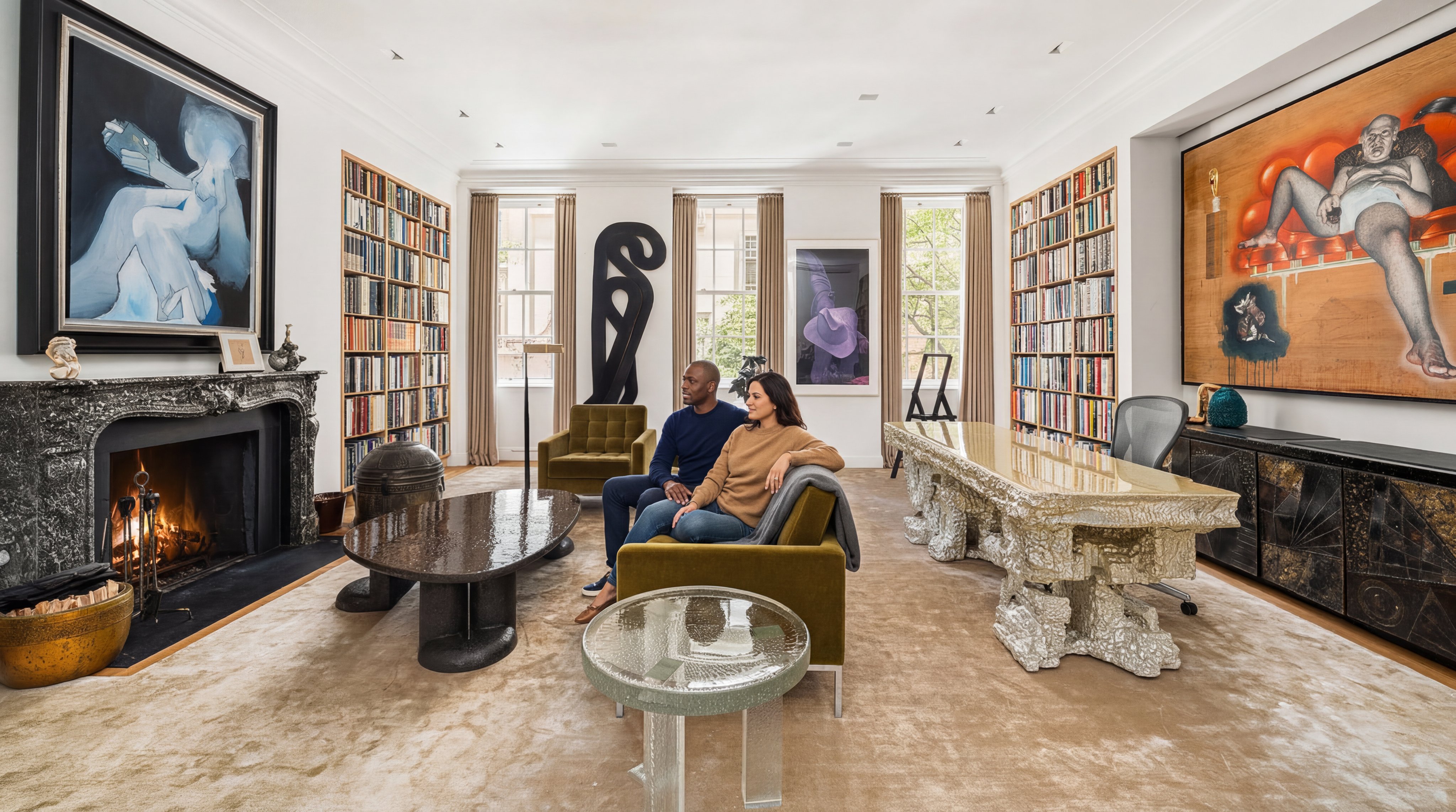 This sophisticated, expansive living room blends formal luxury with a curated gallery aesthetic, featuring high ceilings, dual floor-to-ceiling bookshelves, and a grand marble fireplace. The space is anchored by a velvet sofa and a modernist, sculptural coffee table, complemented by striking, large-scale contemporary artwork. Natural light floods the room through floor-to-ceiling windows, highlighting the luxurious neutral carpeting and high-end design accents.