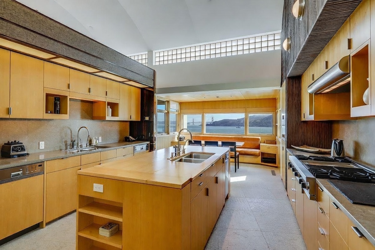 This spacious, modern kitchen features light wood cabinetry, a large central island with a sink, and high-end stainless steel appliances. The room is bathed in natural light from clerestory windows and a large window wall that frames a stunning view of the Golden Gate Bridge. The open-concept design flows seamlessly into a dining area, creating an airy and sophisticated atmosphere.