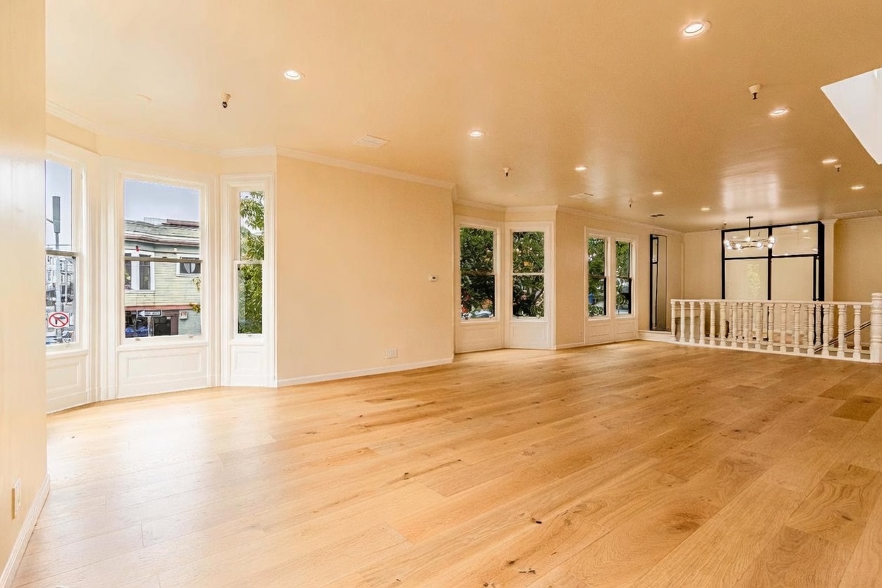 This spacious, open-concept living area features light wood flooring and neutral-toned walls, creating a bright and airy atmosphere. The room is illuminated by numerous recessed ceiling lights and includes multiple large bay windows that provide ample natural light and views of the street. A white wooden railing separates the main living space from a staircase area, adding a classic architectural touch to the modern interior.