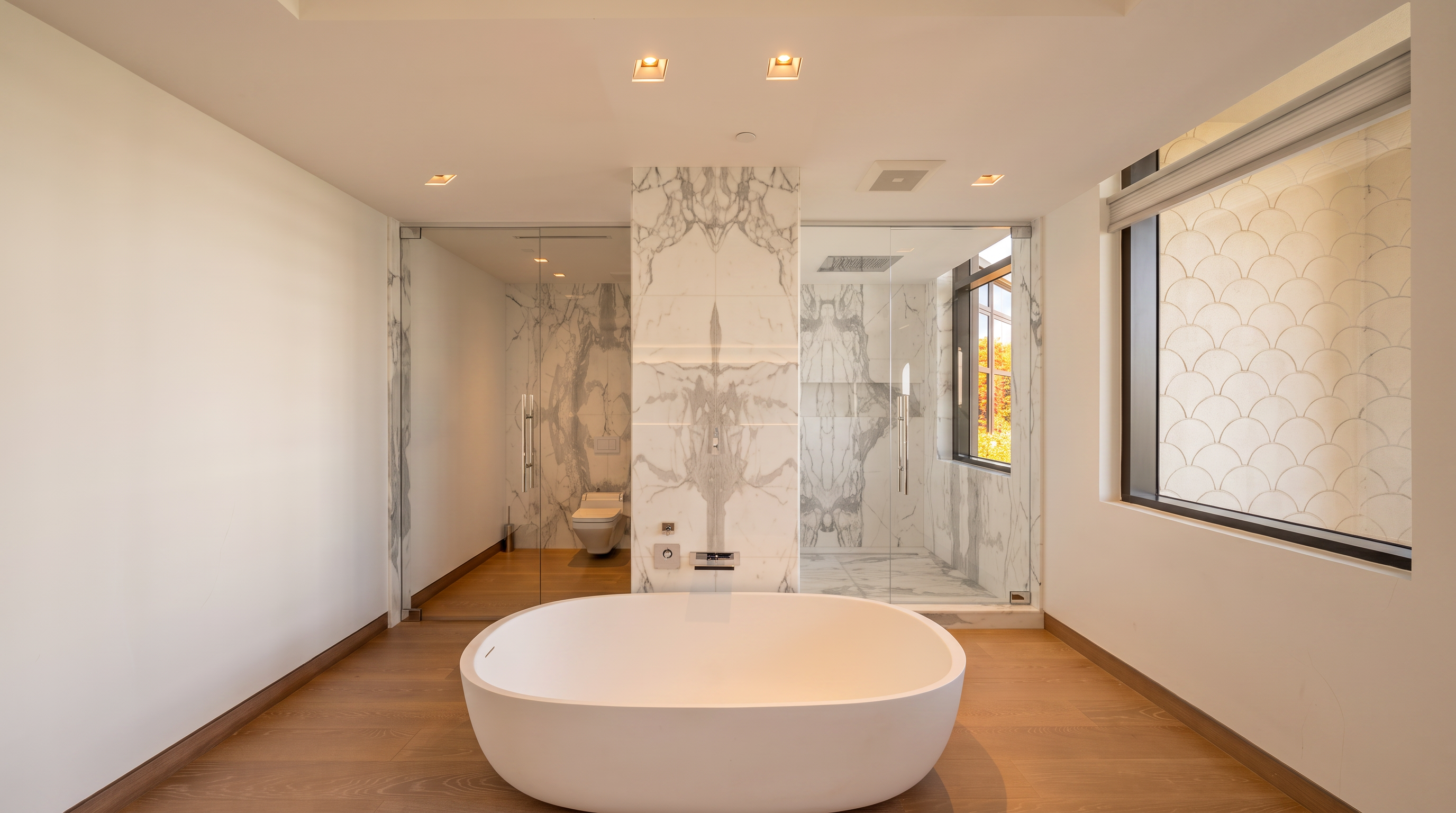 This modern primary bathroom features a central freestanding soaking tub set against a striking stone-clad wall that separates the glass-enclosed shower and toilet area. The room is finished with light wood flooring and crisp white walls, creating a bright and minimalist aesthetic. A large window with patterned tile accents provides natural light, while recessed lighting illuminates the clean, contemporary space.