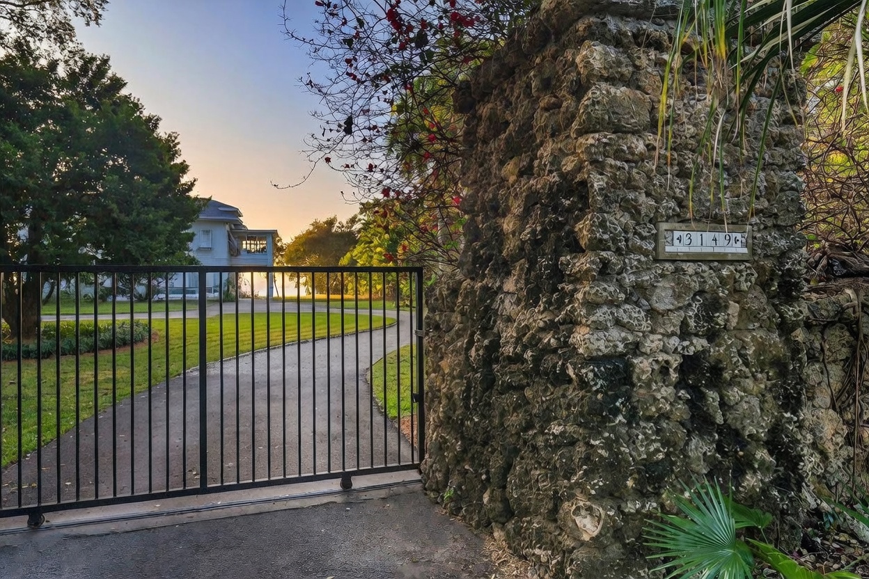 This image captures the gated entrance to a property, featuring a decorative black wrought-iron gate set into a rustic, textured stone pillar. A house number plaque reading '3149' is mounted on the stone wall, which is adorned with climbing vines and foliage. Beyond the gate, a paved driveway leads toward a house partially visible in the distance under a soft, golden-hour sky.