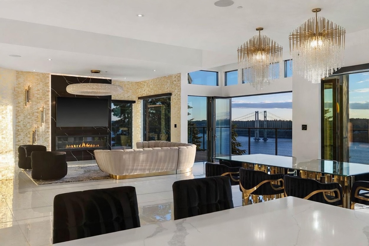 This expansive open-concept living and dining area features a sophisticated, modern aesthetic with polished light-colored flooring and a striking black stone fireplace surround. The space is anchored by a curved, neutral-toned sofa and illuminated by dramatic, tiered crystal chandeliers that add a touch of luxury. Large glass doors open to a scenic view of a bridge over water, seamlessly blending the high-end interior design with the natural landscape.