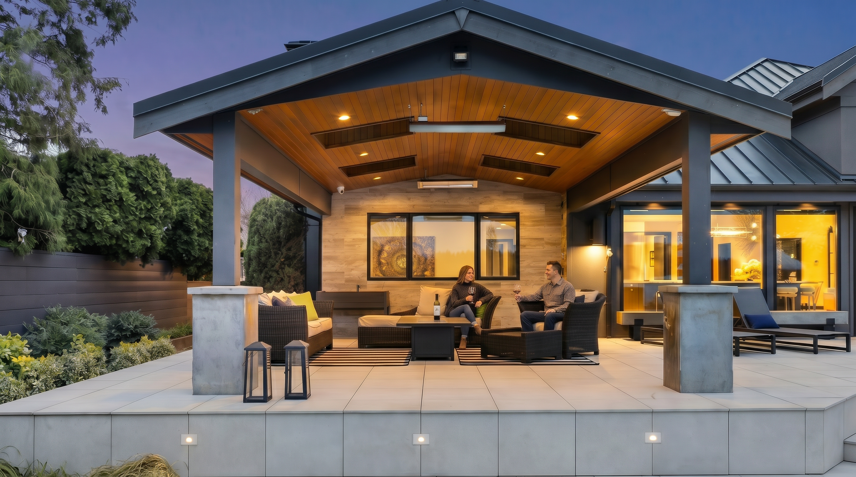 This image showcases a luxurious, covered outdoor patio space designed for sophisticated entertaining. The area features a high-end wood-paneled ceiling with recessed lighting and built-in heaters, a stone-accented wall, and comfortable modern wicker furniture arranged on a striped rug. The perspective is a straight-on, eye-level shot that emphasizes the seamless transition between the indoor living space and the expansive, paved outdoor terrace.
