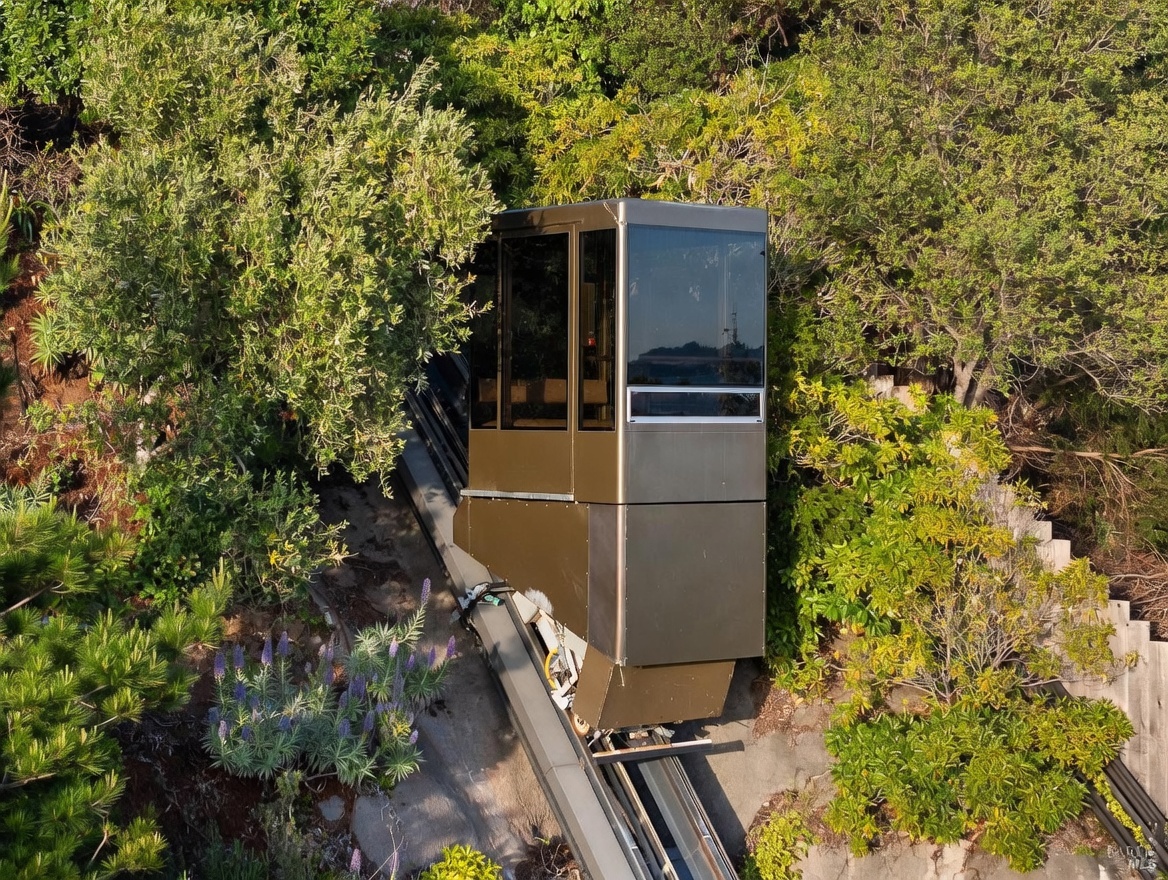 An aerial perspective captures a private funicular or hillside tram car ascending a steep, lushly landscaped slope. The metallic, enclosed cabin features large glass panels, providing a modern and functional solution for navigating challenging terrain. The surrounding foliage is dense and vibrant, emphasizing the secluded and elevated nature of the property.