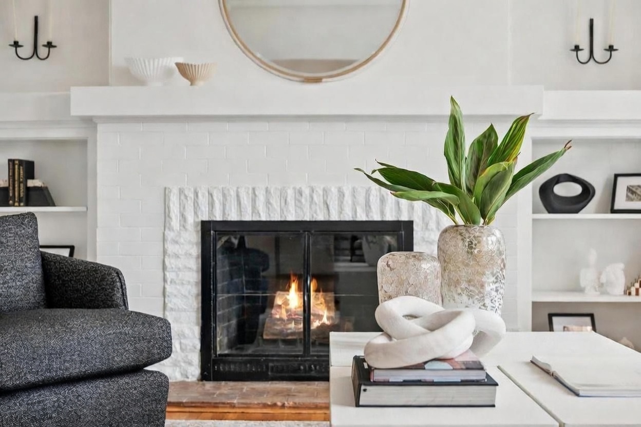 This cozy living room features a white-painted brick fireplace as the central focal point, topped with a simple mantel and a large circular mirror. To the sides, built-in shelving units provide display space, while a dark textured armchair sits in the foreground. A coffee table in the center is styled with books, a sculptural white object, and a glass vase filled with lush green foliage, creating a warm and inviting atmosphere.