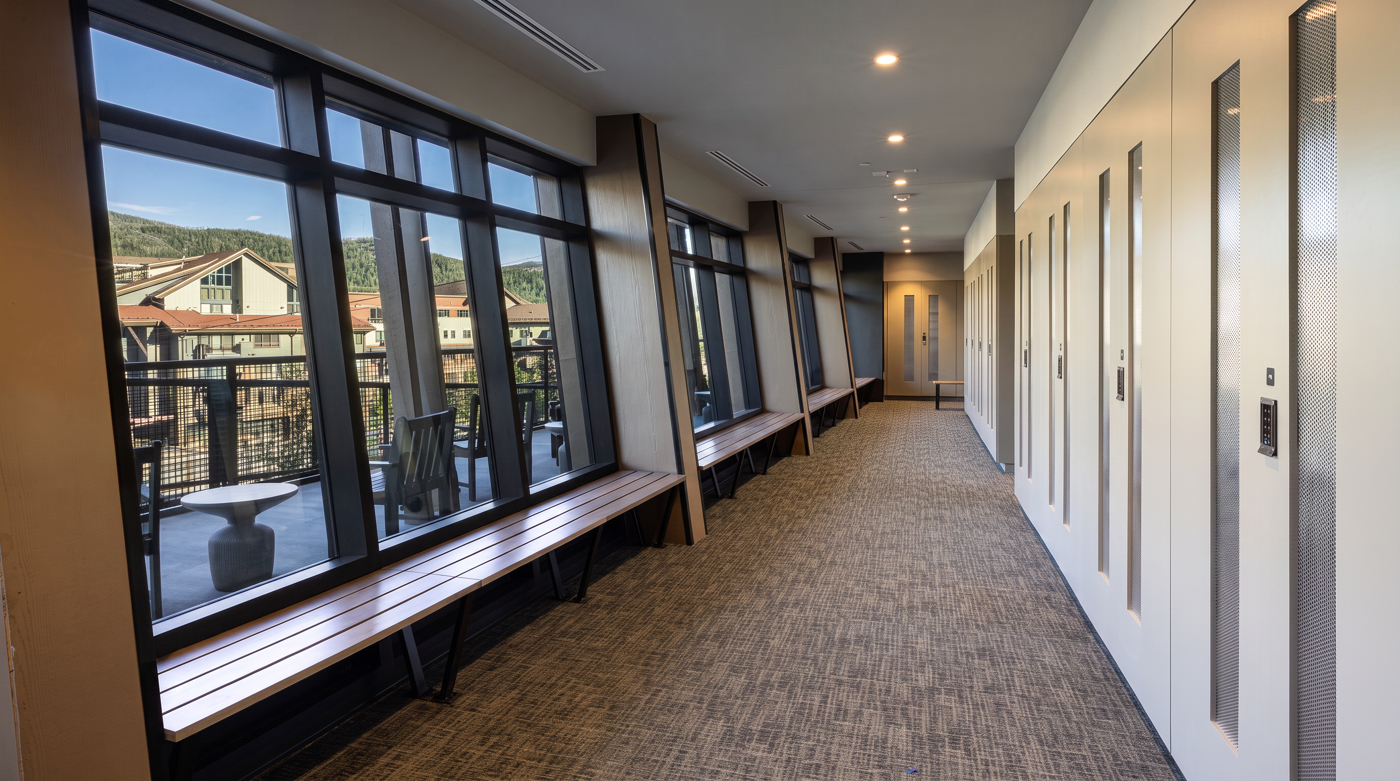 This modern, elongated hallway features a long, built-in wooden bench running along a wall of large windows that offer views of a mountain resort exterior. The opposite wall is lined with a series of tall, light-colored storage lockers with integrated electronic keypads. The perspective draws the eye down the length of the carpeted corridor toward a set of double doors at the far end, creating a clean, functional, and professional atmosphere.