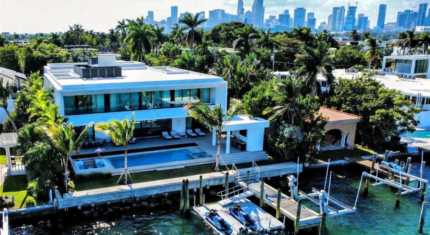This aerial view captures a modern, two-story waterfront residence featuring a sleek, minimalist design with expansive glass walls and a flat roof. The property includes a private swimming pool, a covered outdoor lounge area, and a private dock equipped with jet skis, all set against a lush tropical landscape. In the background, the impressive skyline of a major city provides a dramatic urban contrast to the serene, high-end residential setting.