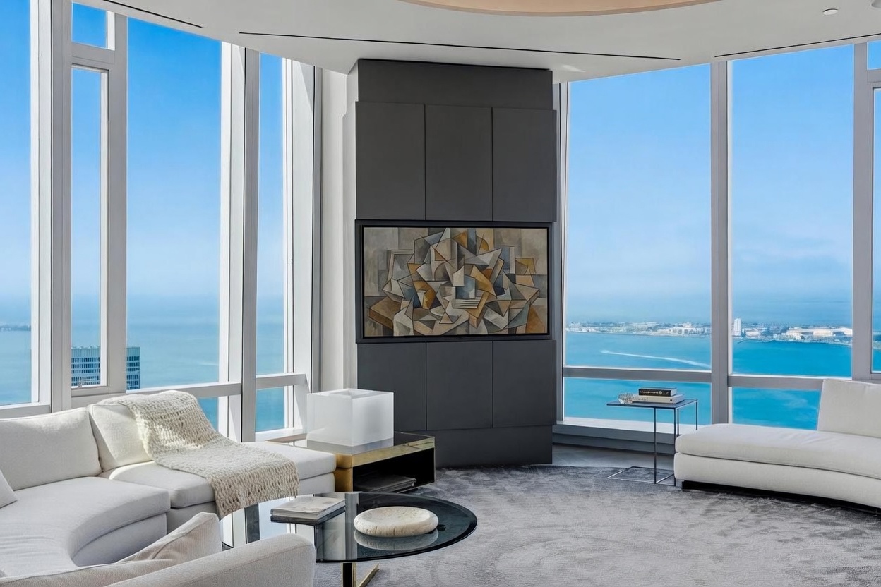 This sophisticated living room features a central, dark-toned column housing a television and artwork, serving as a focal point within the open-concept space. The room is framed by expansive floor-to-ceiling windows that offer panoramic views of the bay and city skyline, bathing the area in natural light. Plush, neutral-toned seating and a circular glass coffee table create an inviting, modern atmosphere perfect for high-end urban living.