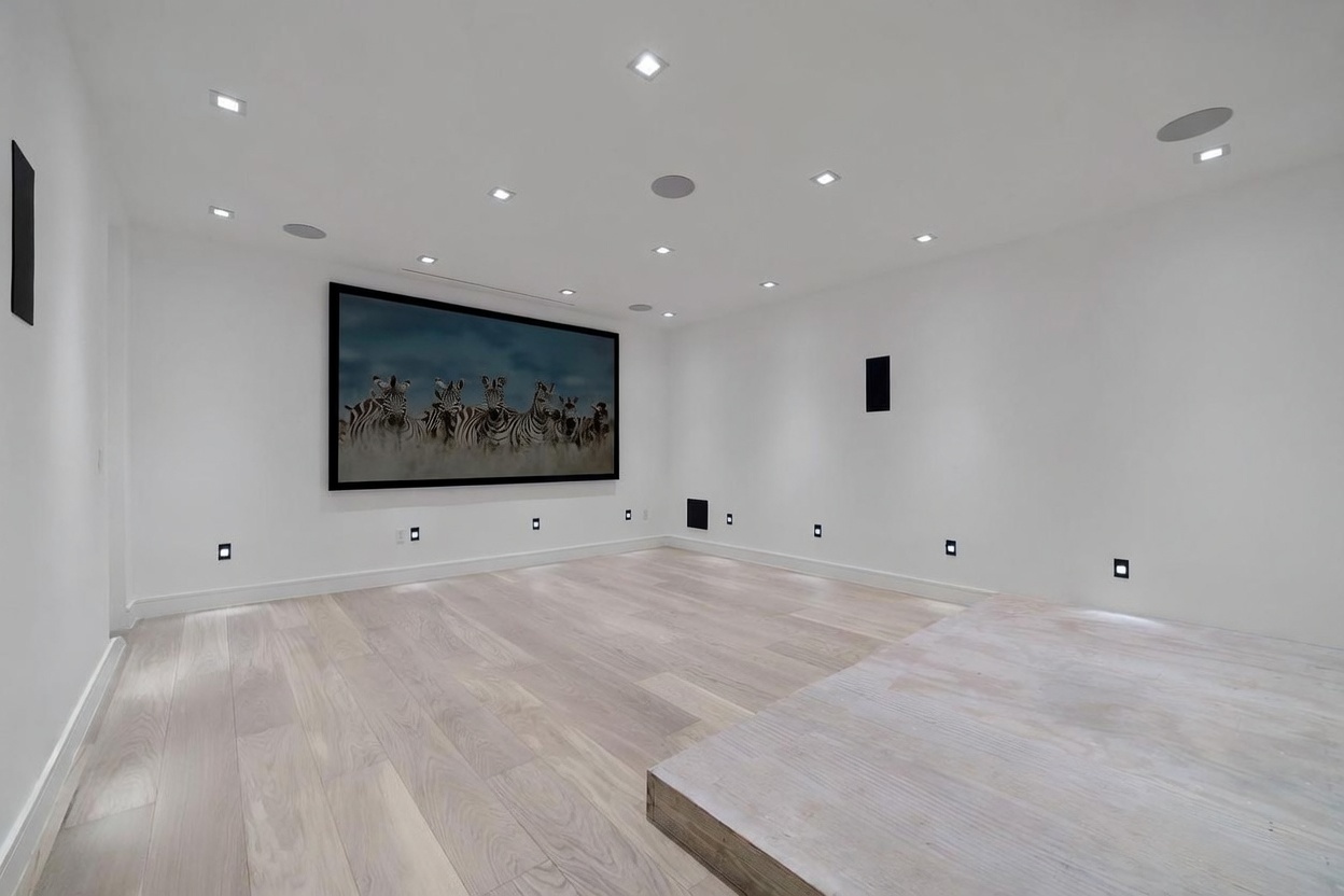 This image showcases a minimalist home theater room featuring light-toned wood flooring and crisp white walls. A large, framed photograph of zebras serves as a focal point on the wall, while recessed lighting and integrated ceiling speakers suggest a high-end media setup. The room includes a raised platform area, providing a tiered layout typical for optimal viewing in a dedicated entertainment space.