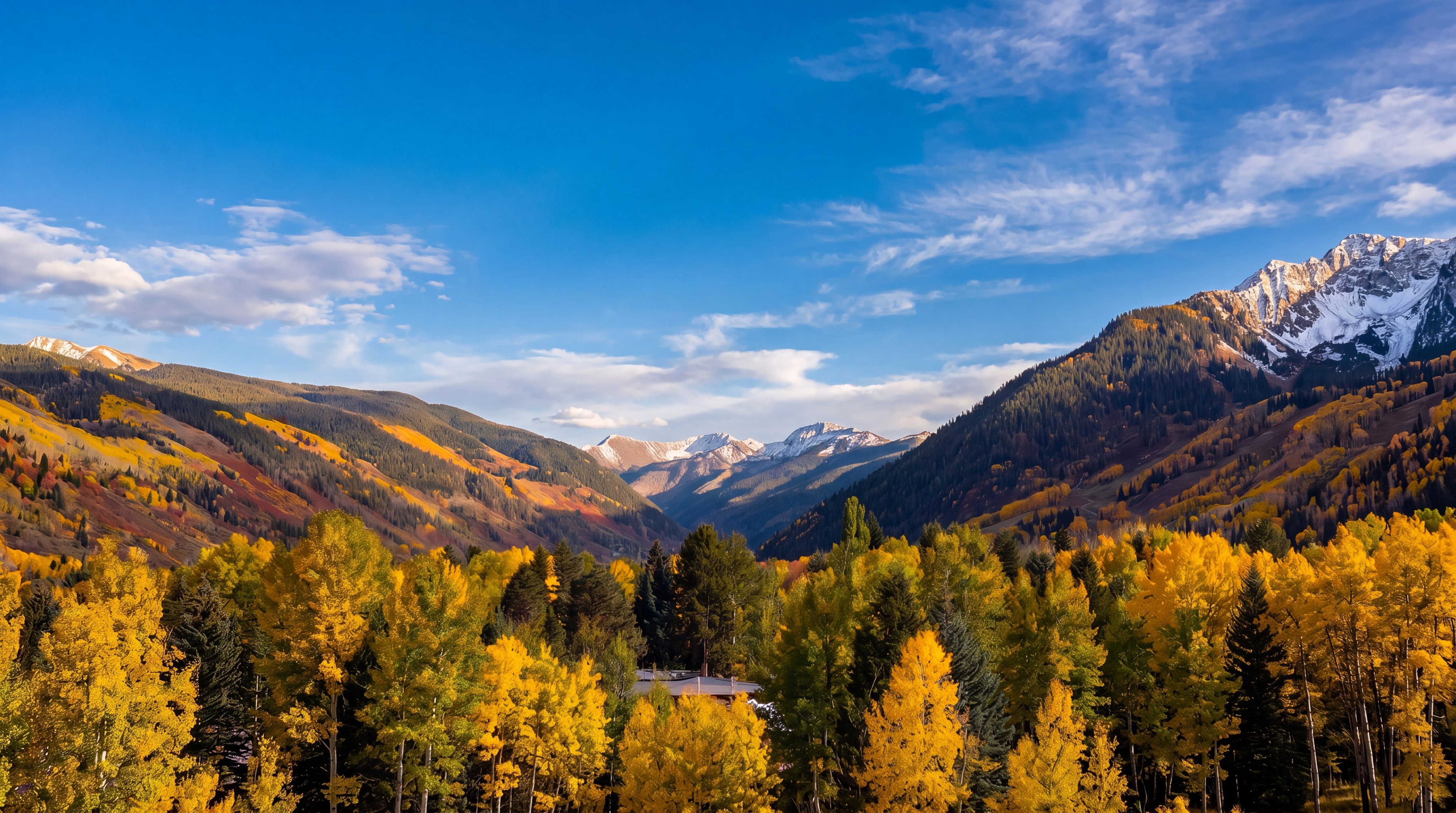 This scenic landscape view captures a breathtaking mountain vista during the autumn season, showcasing vibrant yellow and green foliage in the foreground. The perspective is elevated, looking out over a valley toward rugged, layered mountain peaks under a dramatic sky filled with scattered clouds. The image emphasizes the natural beauty and serene atmosphere of the property's surroundings.