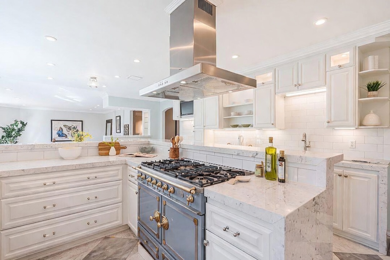 This bright, elegant kitchen features a prominent blue range with gold-toned hardware positioned beneath a stainless steel range hood. The space is defined by white cabinetry, light-colored stone countertops, and a subway tile backsplash that extends across the wall. The perspective captures the kitchen island and cooking area, with a glimpse into an adjacent living space, creating an open and airy atmosphere.