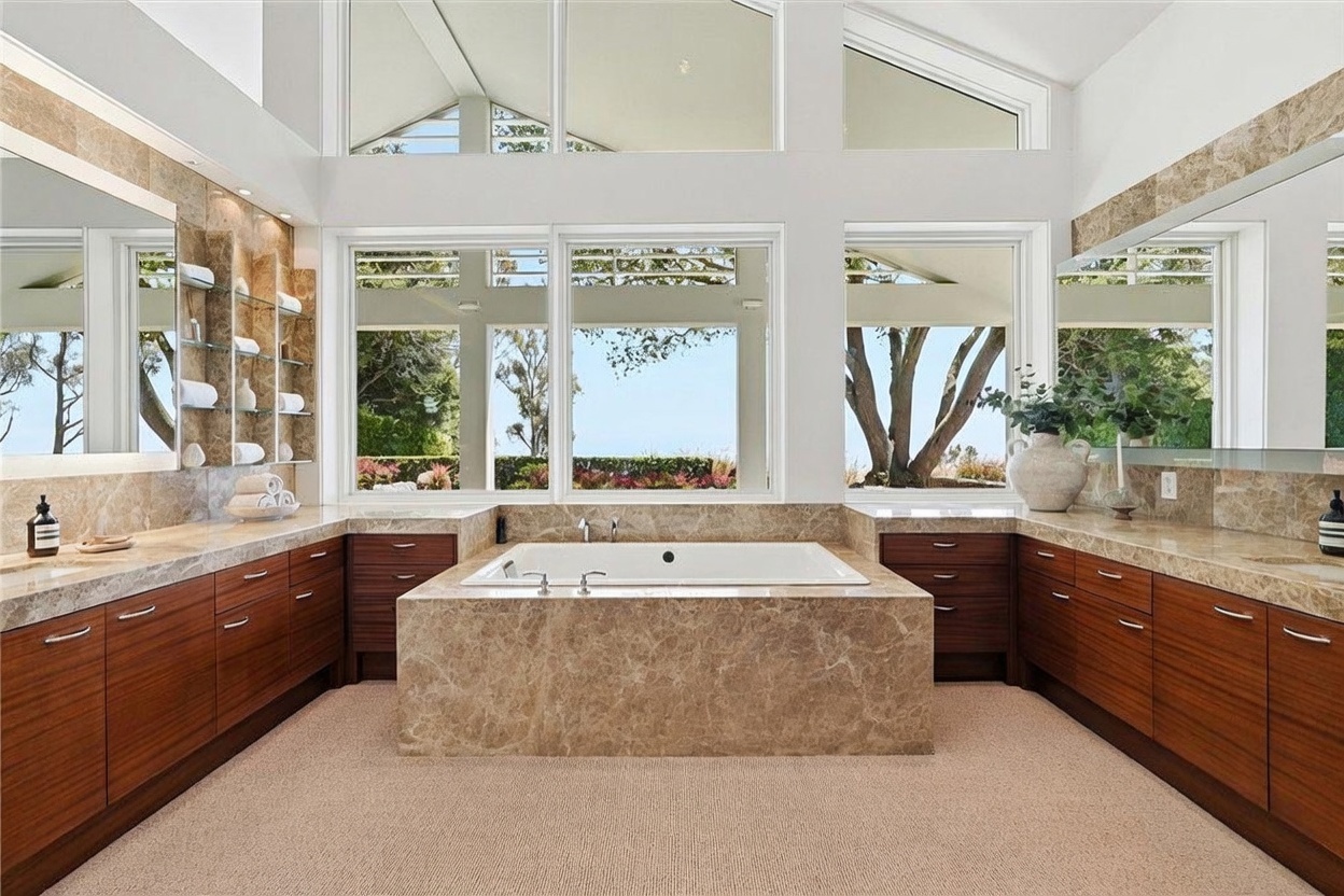 This spacious primary bathroom features a central soaking tub encased in stone, flanked by dual vanities with dark wood cabinetry and stone countertops. Large windows above the tub provide ample natural light and views of the outdoors, while the room is accented by glass shelving and large mirrors. The overall aesthetic is luxurious and serene, with a neutral color palette and high ceilings.