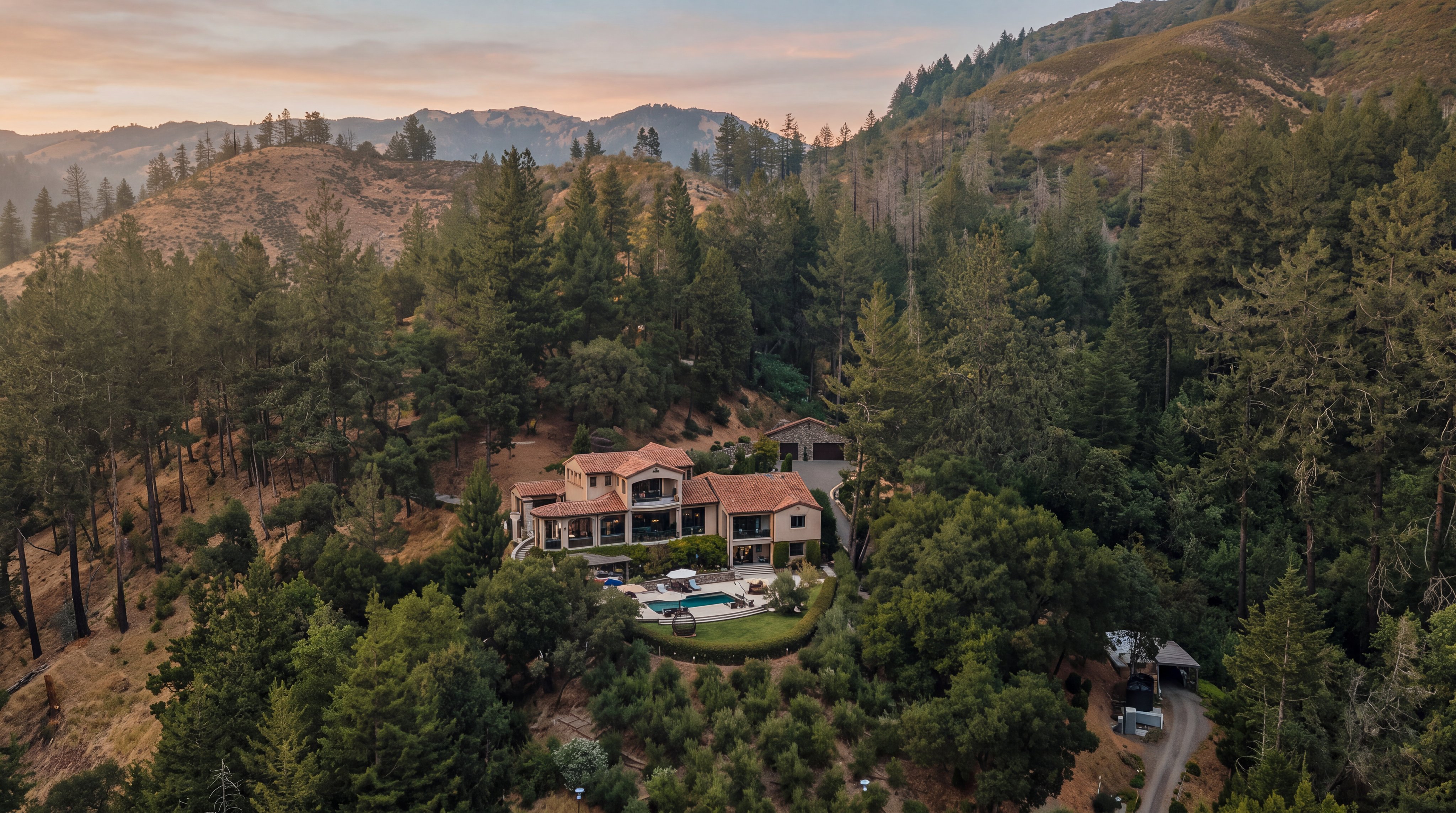 This high-angle aerial view captures a sprawling Mediterranean-style estate nestled within a dense, forested hillside. The property features a multi-level residence with a terracotta tile roof, a private swimming pool set into a landscaped terrace, and a detached garage structure. The surrounding landscape is characterized by lush trees, a winding driveway, and a terraced orchard, creating a secluded and serene mountain retreat.