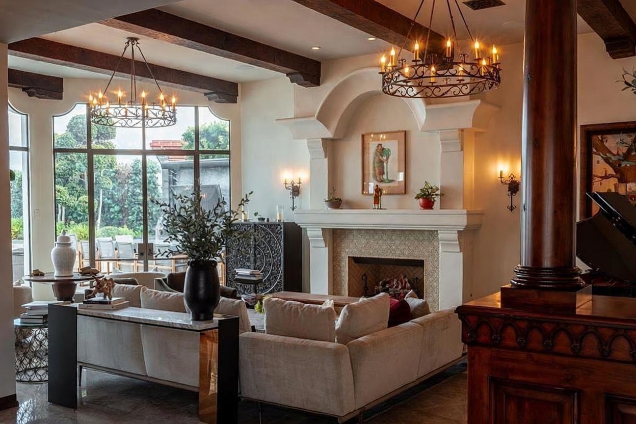 This elegant living room features a prominent fireplace with a decorative arched surround and a tiled hearth, serving as the focal point of the space. The room is anchored by a large, light-colored sectional sofa and a console table, complemented by exposed dark wood ceiling beams and two ornate iron chandeliers. Large windows provide ample natural light and a view of the outdoor greenery, creating a sophisticated and inviting atmosphere.