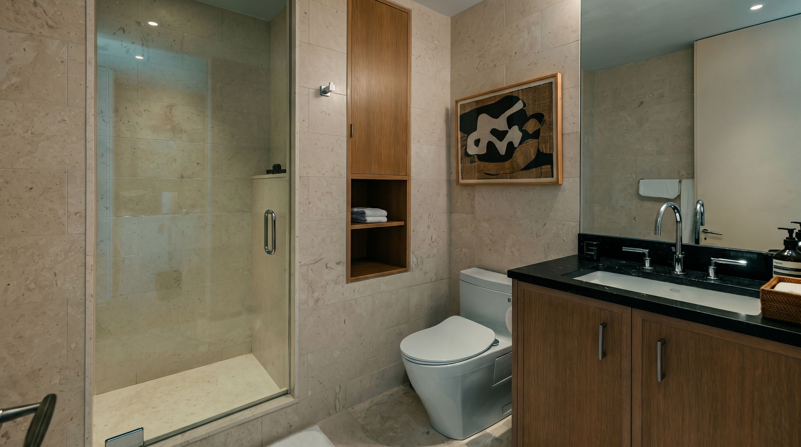 This modern bathroom features a glass-enclosed shower with light-toned stone wall tiles and a matching stone floor. A vanity with a dark countertop and light wood cabinetry sits beside a white toilet, while a built-in shelving unit provides convenient storage. The space is brightened by a large mirror and a framed piece of abstract art, creating a clean and contemporary aesthetic.