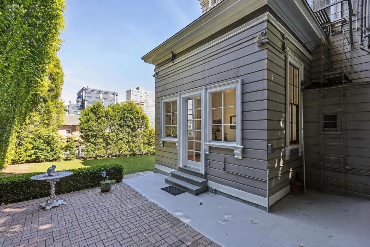 This exterior view captures a private brick-paved patio area adjacent to a gray-sided home. The space features a glass-paned door leading into the residence, flanked by two windows, and includes a small decorative metal table. A lush green lawn and mature trees provide a serene backdrop, creating a quiet and inviting outdoor living space.