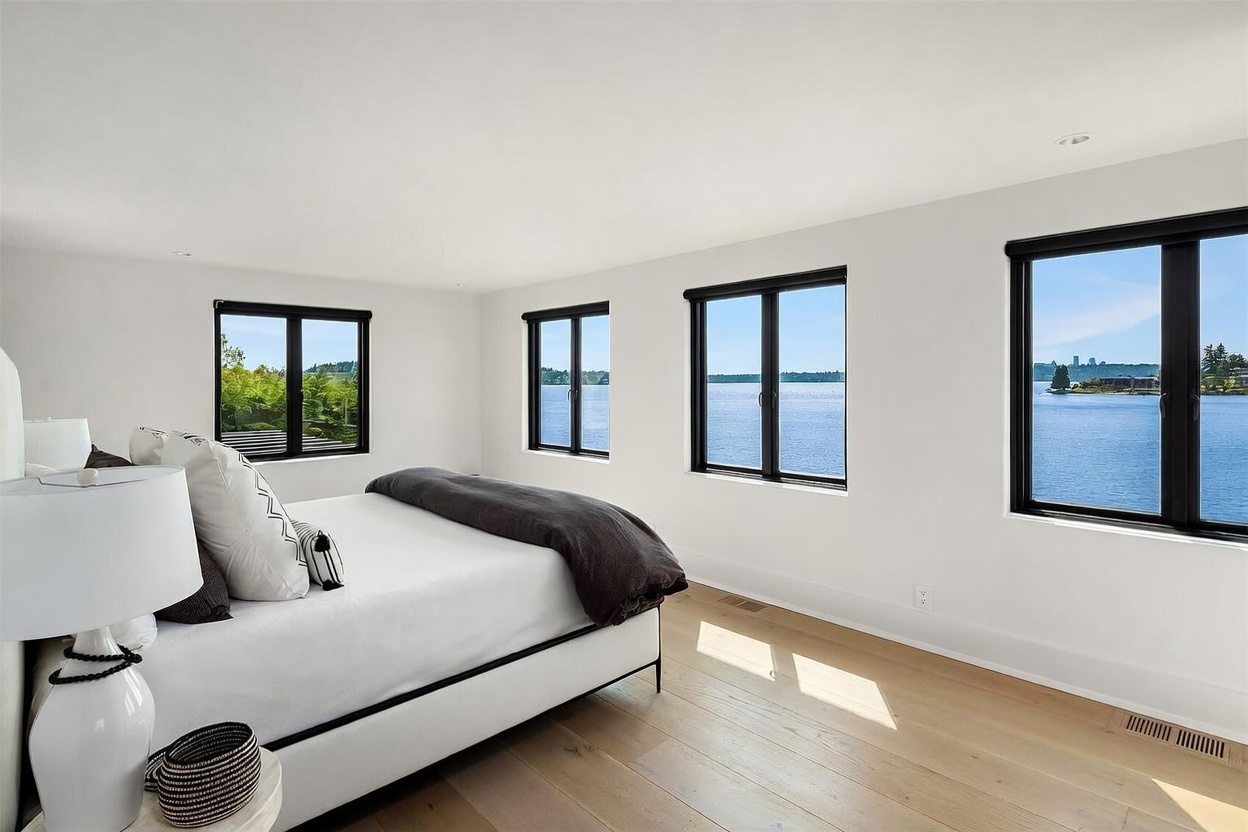 This bright and airy primary bedroom features light wood flooring and a minimalist aesthetic with white walls. A bed with a dark-toned throw blanket is positioned to take advantage of the expansive water views provided by the multiple large windows lining the wall. The room offers a serene, tranquil atmosphere with a clean, modern design that emphasizes the natural scenery outside.
