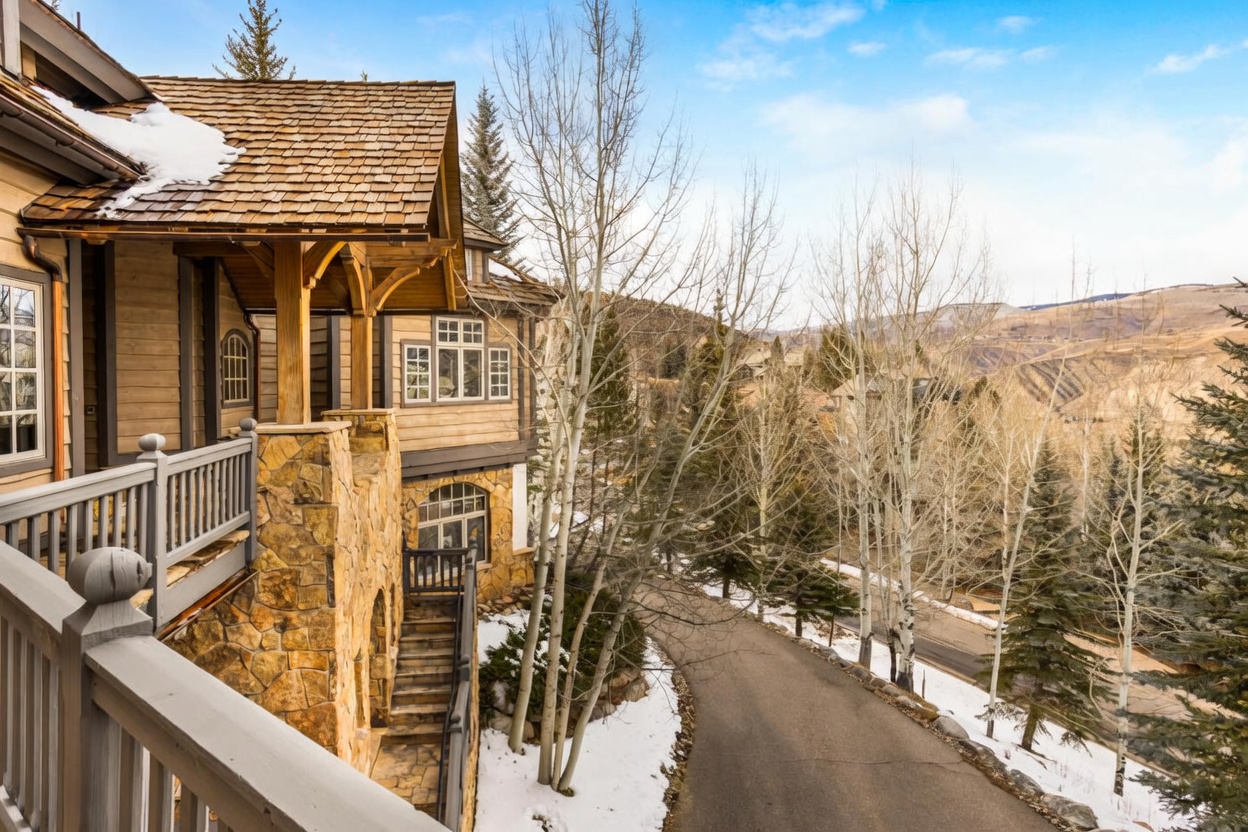 This side view captures a rustic mountain home featuring a combination of wood siding and stone masonry. A wooden balcony with a railing overlooks a winding paved driveway and a wooded landscape with tall, slender trees. The scene is set against a backdrop of rolling hills under a bright, partly cloudy sky, emphasizing the property's elevated and scenic location.