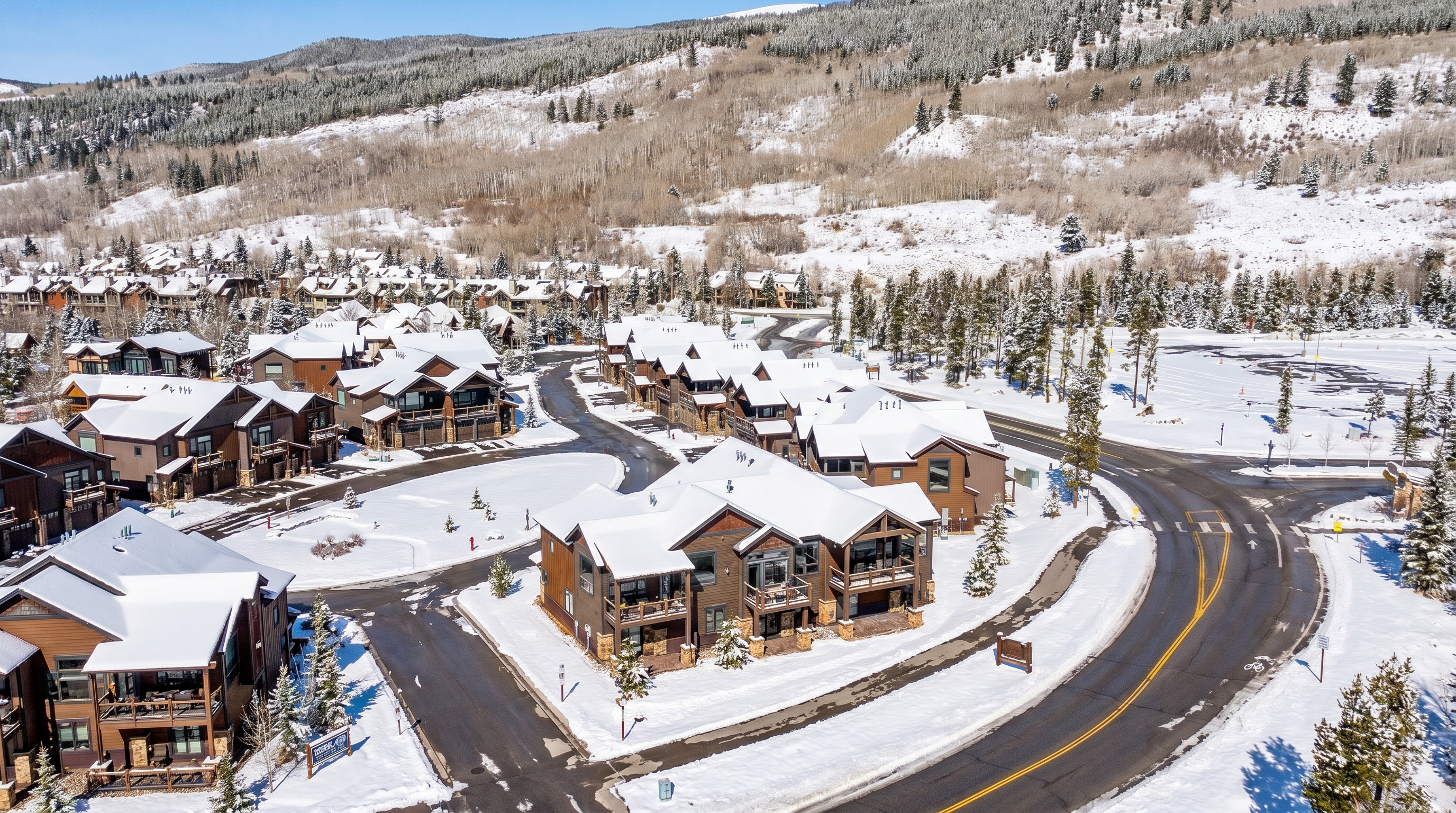 This high-angle aerial shot captures a modern mountain residential community nestled at the base of a vast, forested hillside. The scene features several multi-story townhomes with distinctive dark shingled roofs and rustic metal accents, connected by winding paved roads and landscaped pathways. The bright, clear blue sky and expansive natural backdrop emphasize the serene, upscale nature of this mountain retreat.