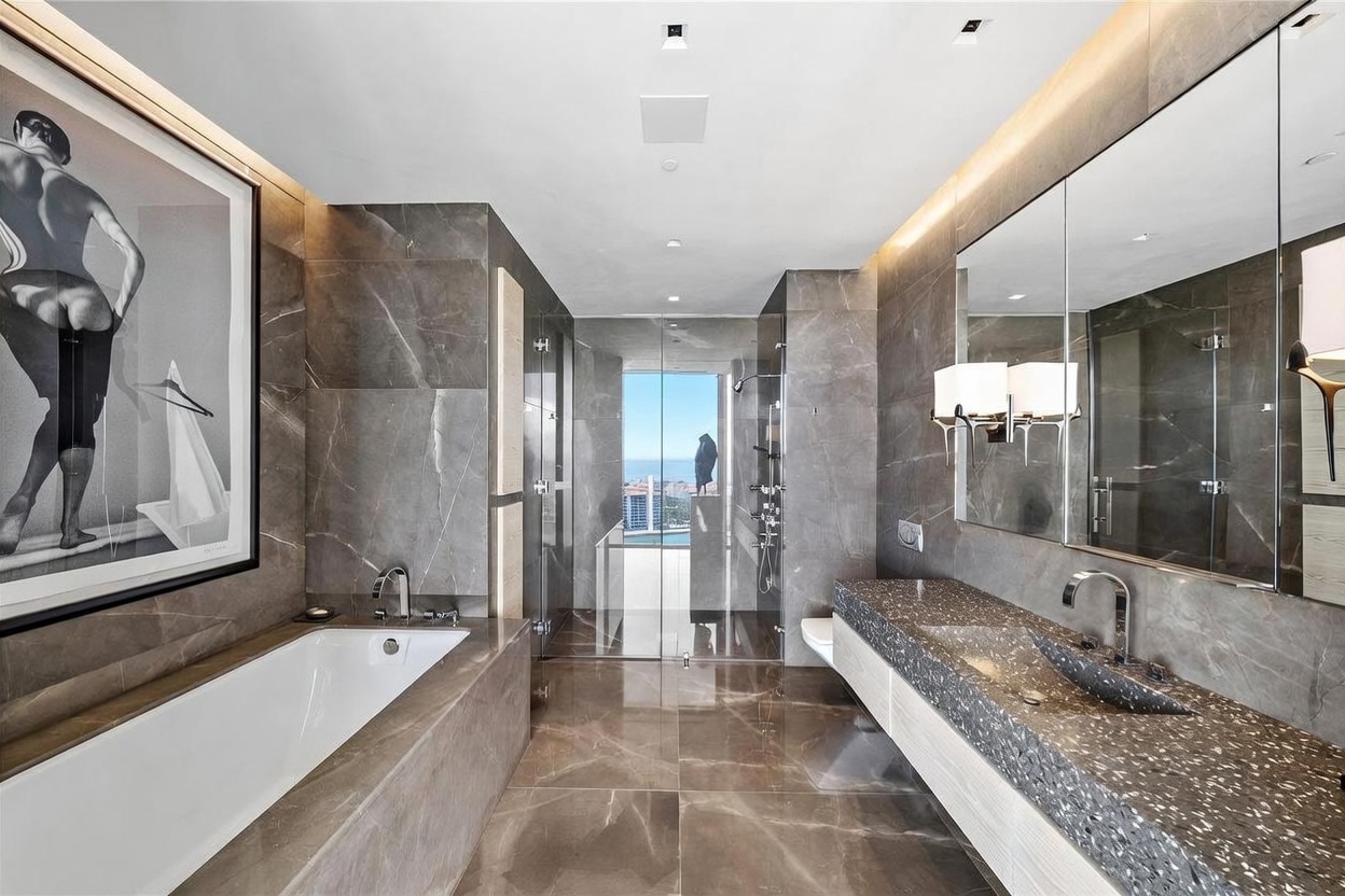 This luxurious primary bathroom features dark, large-format stone tiling throughout the walls and floor, creating a sophisticated and moody aesthetic. The space includes a deep soaking tub, a floating vanity with a textured stone countertop, and a glass-enclosed shower that offers a clear view of the city skyline through a large window. A large framed art piece adds a personal touch, while integrated lighting enhances the modern, high-end atmosphere.