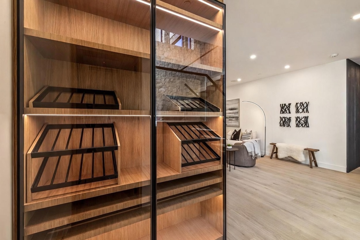 This image features a sophisticated, modern wine storage unit with glass doors and integrated LED lighting, showcasing wooden display racks for wine bottles. The perspective looks through the glass shelving into an adjacent living area, which is furnished with a comfortable armchair and a decorative bench. The space is characterized by a clean, contemporary aesthetic with light wood flooring and a textured stone wall visible in the background.