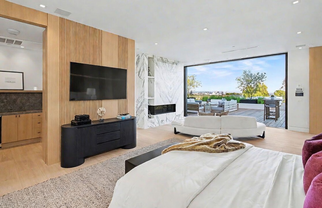 This spacious primary bedroom features a contemporary design with light wood flooring and a textured wood-slat accent wall behind the television. A large white curved sofa sits near the expansive glass wall that opens onto a private deck, providing a seamless transition to the outdoors. The room is anchored by a stone-clad chimney breast and a plush bed, creating a luxurious and serene atmosphere.