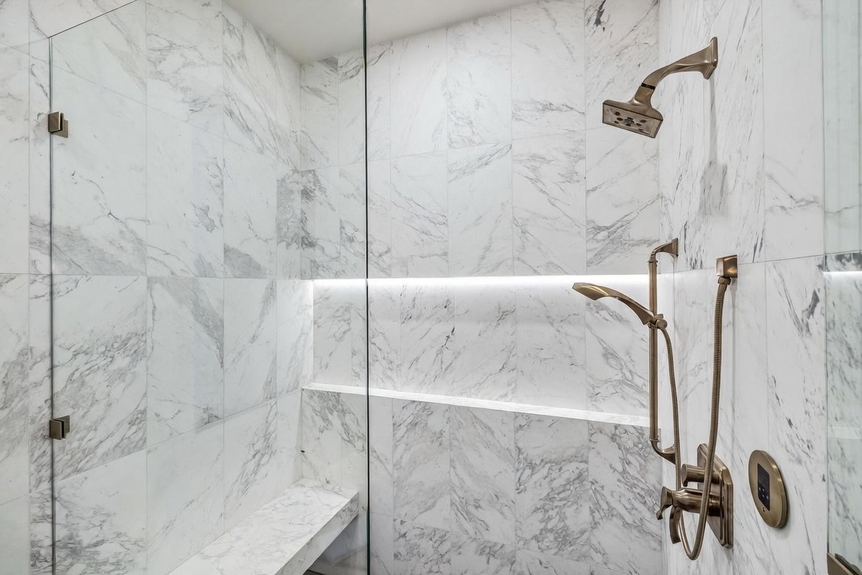 This modern walk-in shower features large-format, light-colored stone tiles with a distinct grey veining pattern throughout the walls. A built-in bench provides seating, while a recessed niche with integrated LED lighting adds a sophisticated, functional accent. The shower is equipped with a wall-mounted showerhead and a separate handheld sprayer, both finished in a warm metallic tone, and is enclosed by a clear glass panel.
