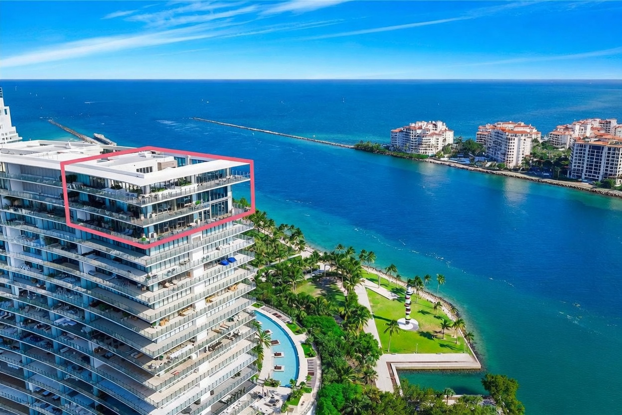 This high-angle aerial view captures a modern luxury high-rise building situated along a vibrant blue waterway. The image highlights the building's expansive balconies and a prominent rooftop terrace, while the surrounding landscape features a lush green park, a swimming pool, and a scenic jetty extending into the ocean. The perspective emphasizes the property's prime waterfront location and its integration with the coastal environment.