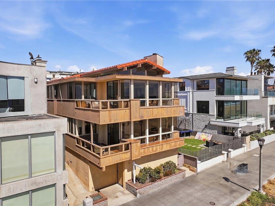This elevated perspective captures a multi-story coastal residence featuring a distinctive wood-paneled facade and a terracotta-tiled roof. The home includes multiple levels of balconies with glass railings, offering expansive views, while a small, manicured lawn sits adjacent to the structure. The scene is set in a bright, sunny environment, highlighting the property's proximity to the beach and its unique architectural design.
