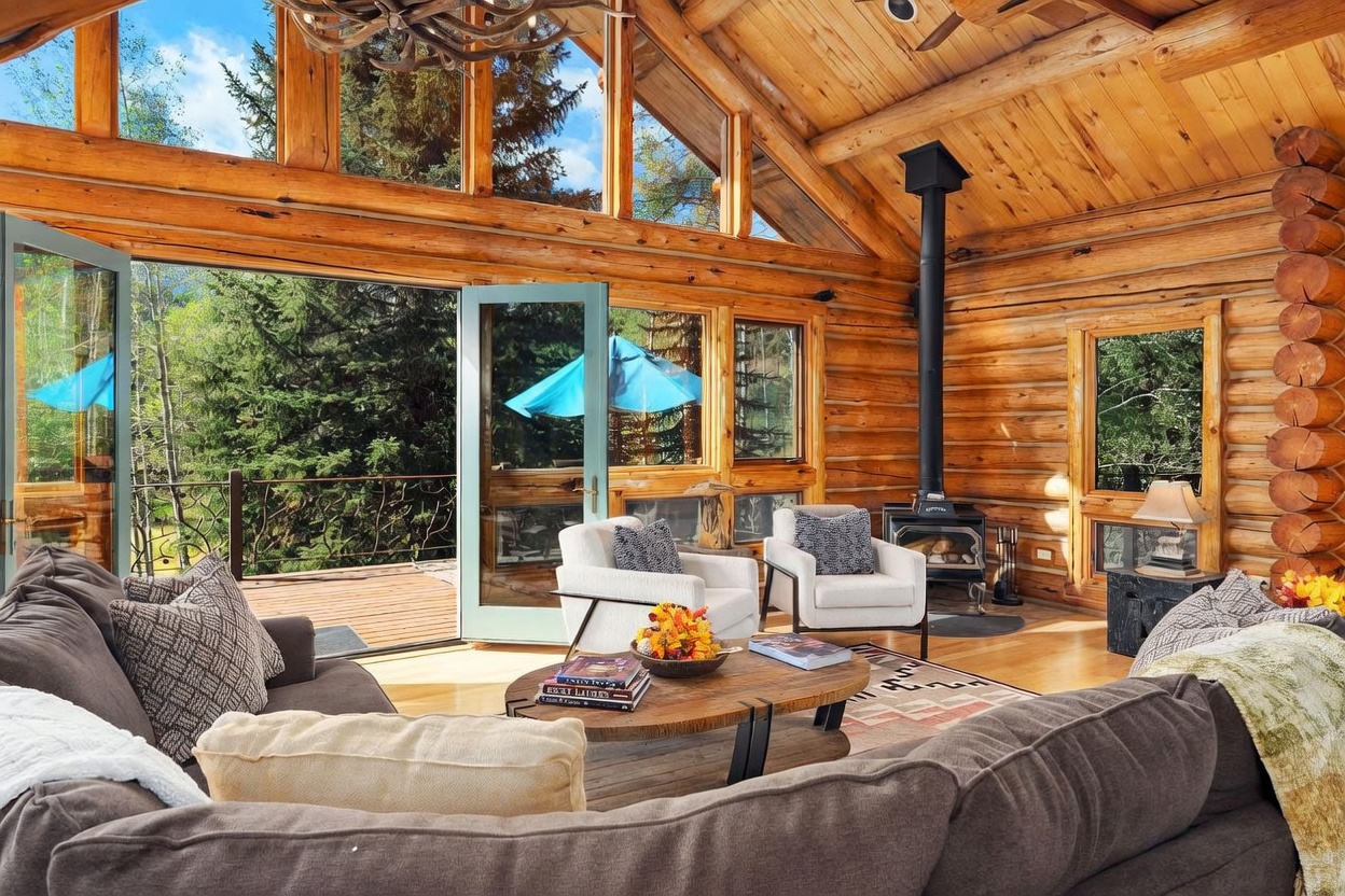 This rustic living room features exposed log walls, a vaulted wood-paneled ceiling, and large windows that provide an expansive view of the surrounding forest. A central wood-burning stove stands on a hearth, while a comfortable seating area with a plush sofa and armchairs is arranged around a circular wooden coffee table. The open-concept design flows seamlessly onto an outdoor deck through wide-opening glass doors, creating a bright and inviting atmosphere.