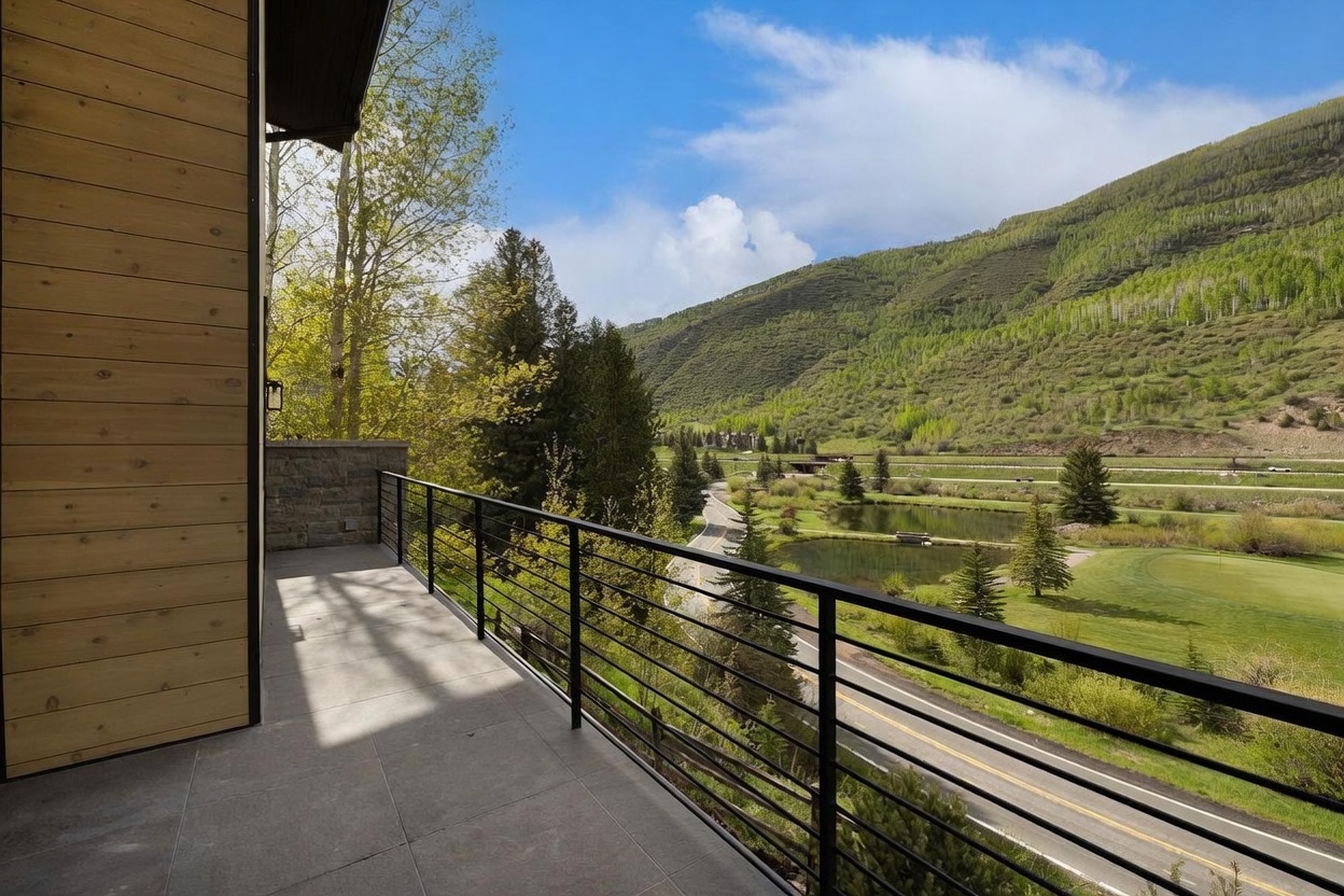 This elevated balcony features light-toned wood siding on the wall and a clean, tiled floor surface. A sleek black metal railing provides safety while maintaining an unobstructed view of the surrounding landscape, which includes a winding road, small ponds, and lush green hills. The perspective captures a serene, expansive outdoor living space perfect for enjoying the natural scenery.
