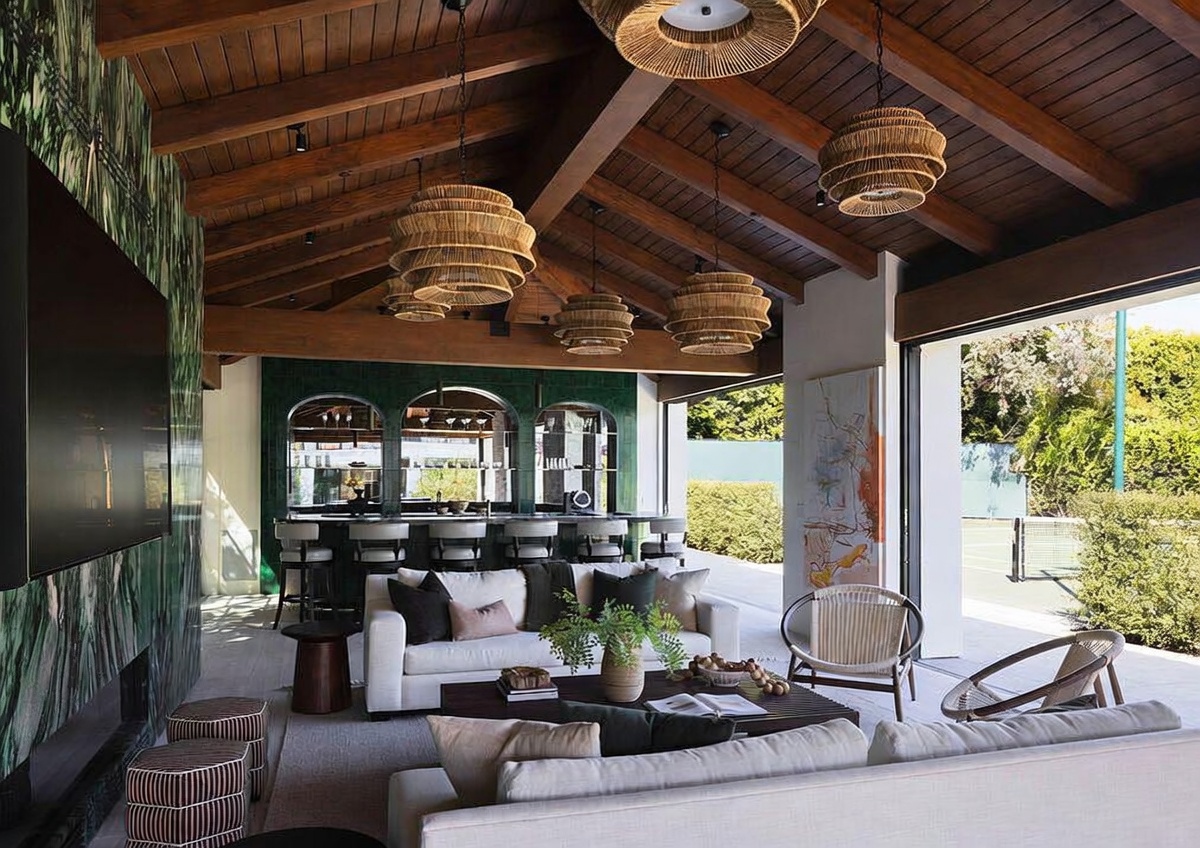 This expansive, open-air living space features a vaulted wood-paneled ceiling with exposed beams and multiple woven pendant lights. The room transitions seamlessly to an outdoor area, showcasing a bar with arched mirrors and a comfortable seating arrangement centered around a coffee table. The aesthetic is sophisticated and airy, blending indoor comfort with an outdoor lifestyle.