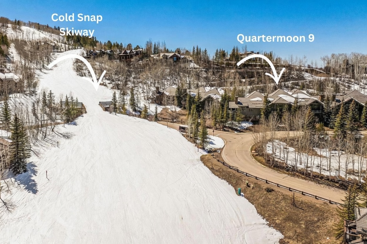 This aerial perspective captures a scenic mountain residential area nestled against a snowy ski slope. The image highlights the proximity of luxury homes to the 'Cold Snap' skiway and 'Quartermoon 9' trail, showcasing the prime ski-in/ski-out location. A winding paved road curves through the wooded landscape, connecting the properties amidst a backdrop of clear blue skies and evergreen trees.