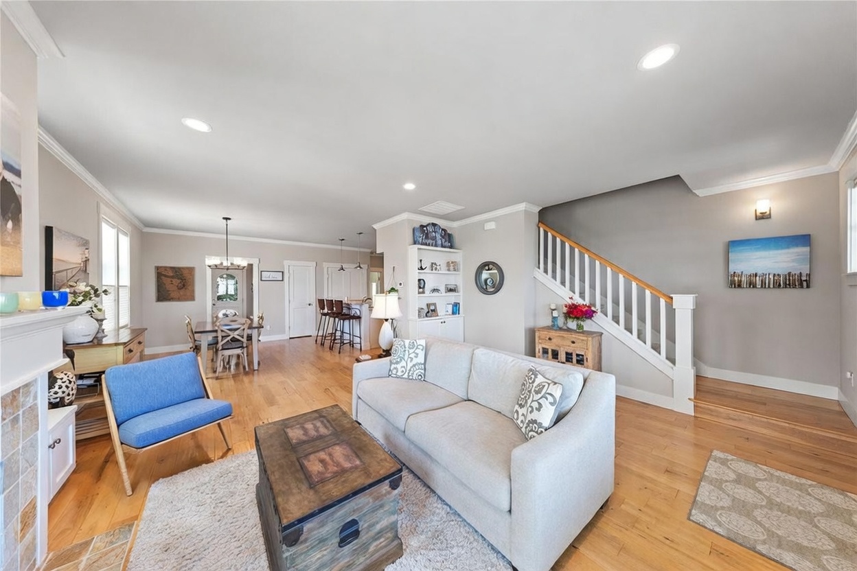 This open-concept living area features light wood flooring and a neutral color palette, creating a bright and inviting atmosphere. The space includes a comfortable sofa, a rustic wooden coffee table, and a vibrant blue accent chair, with a staircase leading to the upper level in the background. The view extends into the dining area and kitchen, showcasing a cohesive flow perfect for modern living.