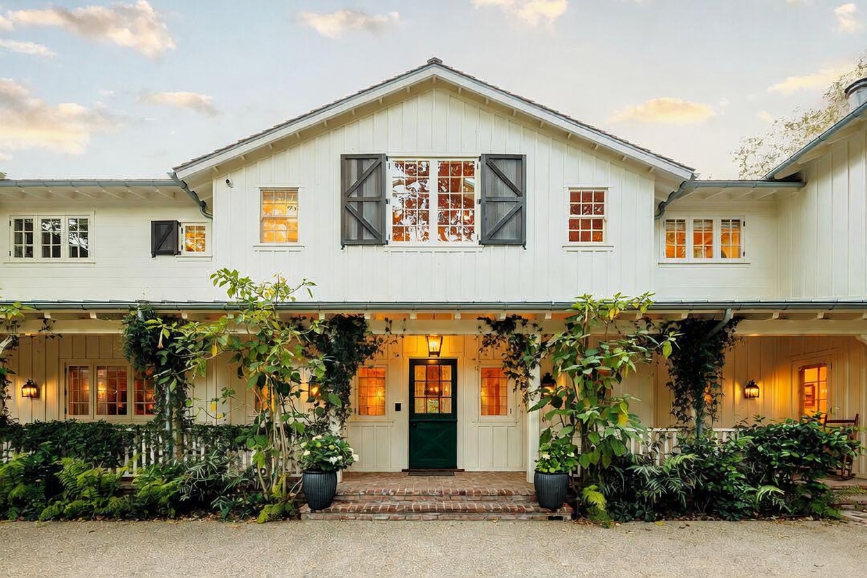 This charming farmhouse-style home features a symmetrical white board-and-batten facade with a welcoming covered porch spanning the front. The exterior is accented by dark shutters, a central green door, and lush greenery that softens the architectural lines. The warm glow from the interior lights and exterior lanterns creates an inviting and serene atmosphere at dusk.