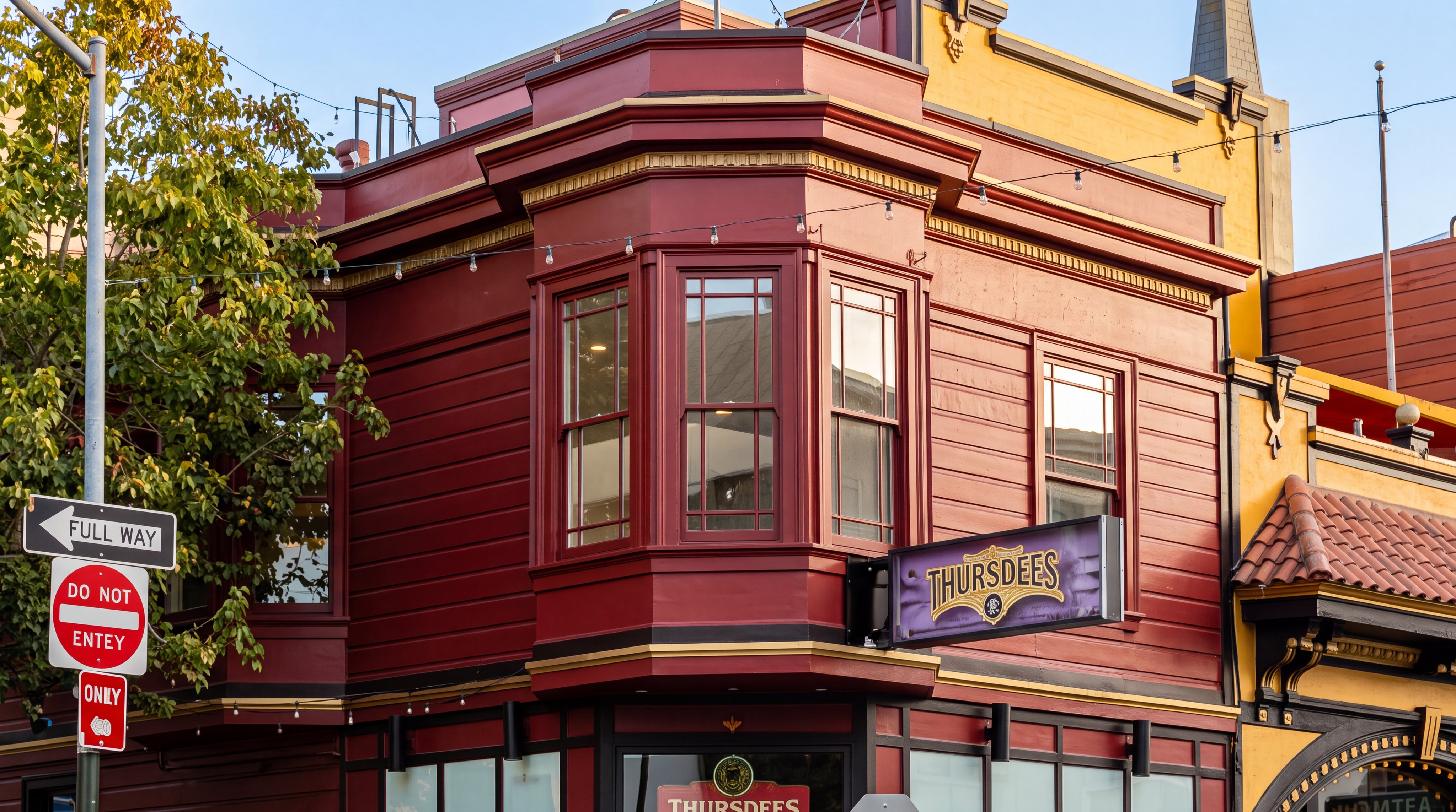 This building features a distinct architectural style with deep red wooden siding, prominent bay windows, and gilded trim accents. The structure serves as a commercial location, indicated by the 'THURSDEES' signage and storefront windows. Its classic design and vibrant color make it a striking landmark on the corner.