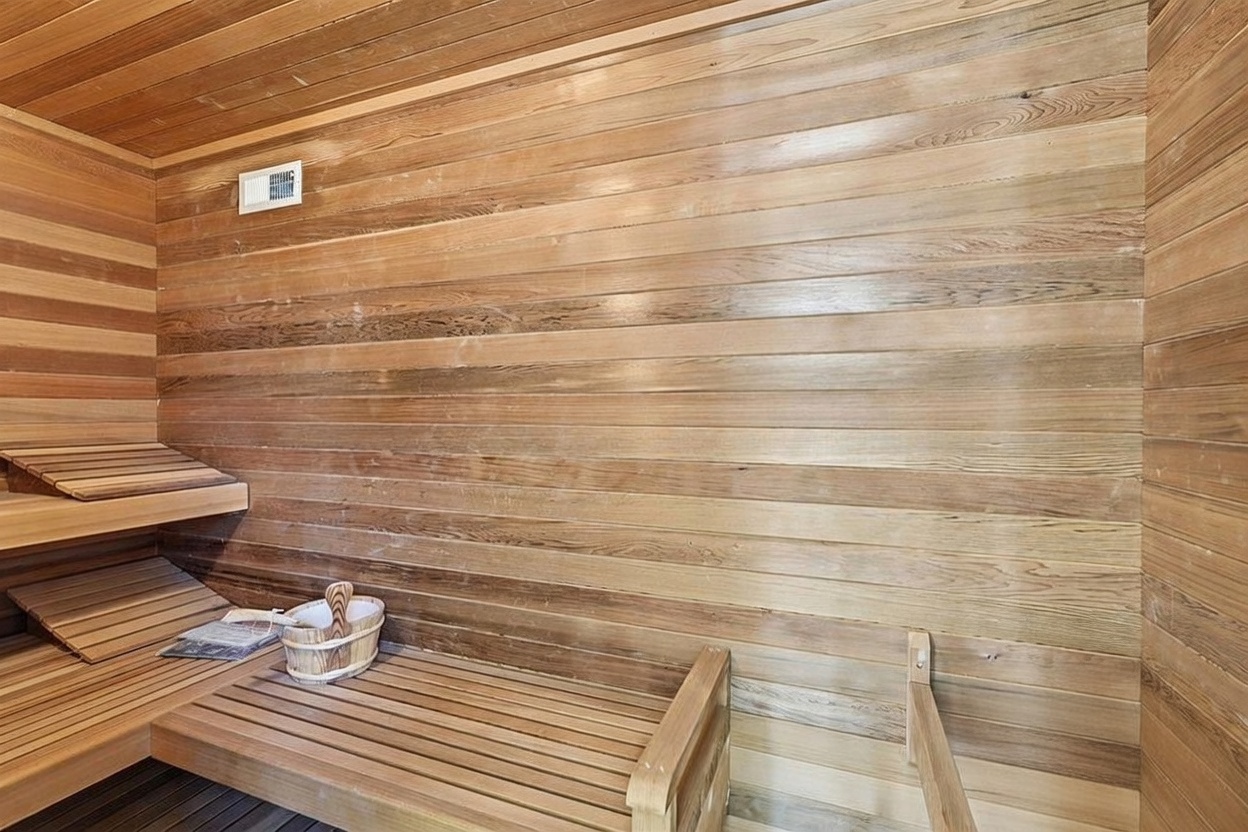 This image showcases a traditional wood-paneled sauna, featuring tiered benches and a dedicated heater filled with volcanic rocks. The warm, natural wood tones create a cozy and relaxing atmosphere, typical of a high-end home spa amenity. The perspective is from inside the sauna, looking toward the corner where the heater is positioned.