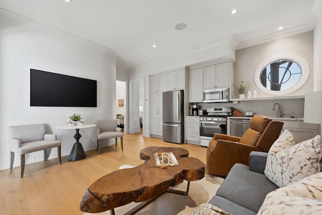 This open-concept living space features a blend of modern and rustic elements, including a unique live-edge wood coffee table and a cozy leather recliner. The room flows seamlessly into a compact kitchen area equipped with stainless steel appliances and light-toned cabinetry. A large wall-mounted television and a small dining nook with two chairs complete the functional layout, all set against a backdrop of light wood flooring and a distinctive circular window.