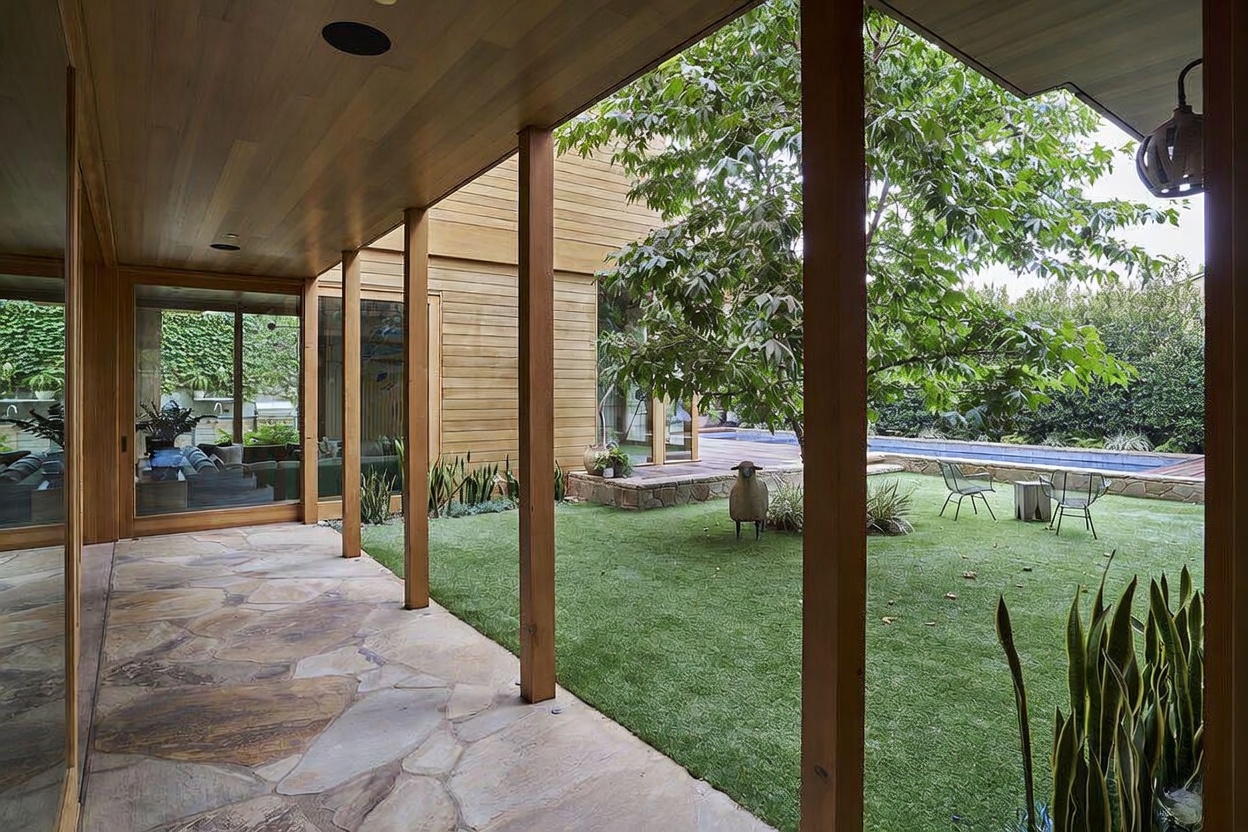 This image captures a covered stone patio that transitions seamlessly into a lush, manicured lawn and a private swimming pool area. The space features warm wood-paneled ceilings and structural posts, creating a sophisticated indoor-outdoor flow through large glass walls. A whimsical sheep sculpture adds a touch of character to the serene backyard, which is framed by mature trees and dense perimeter greenery.