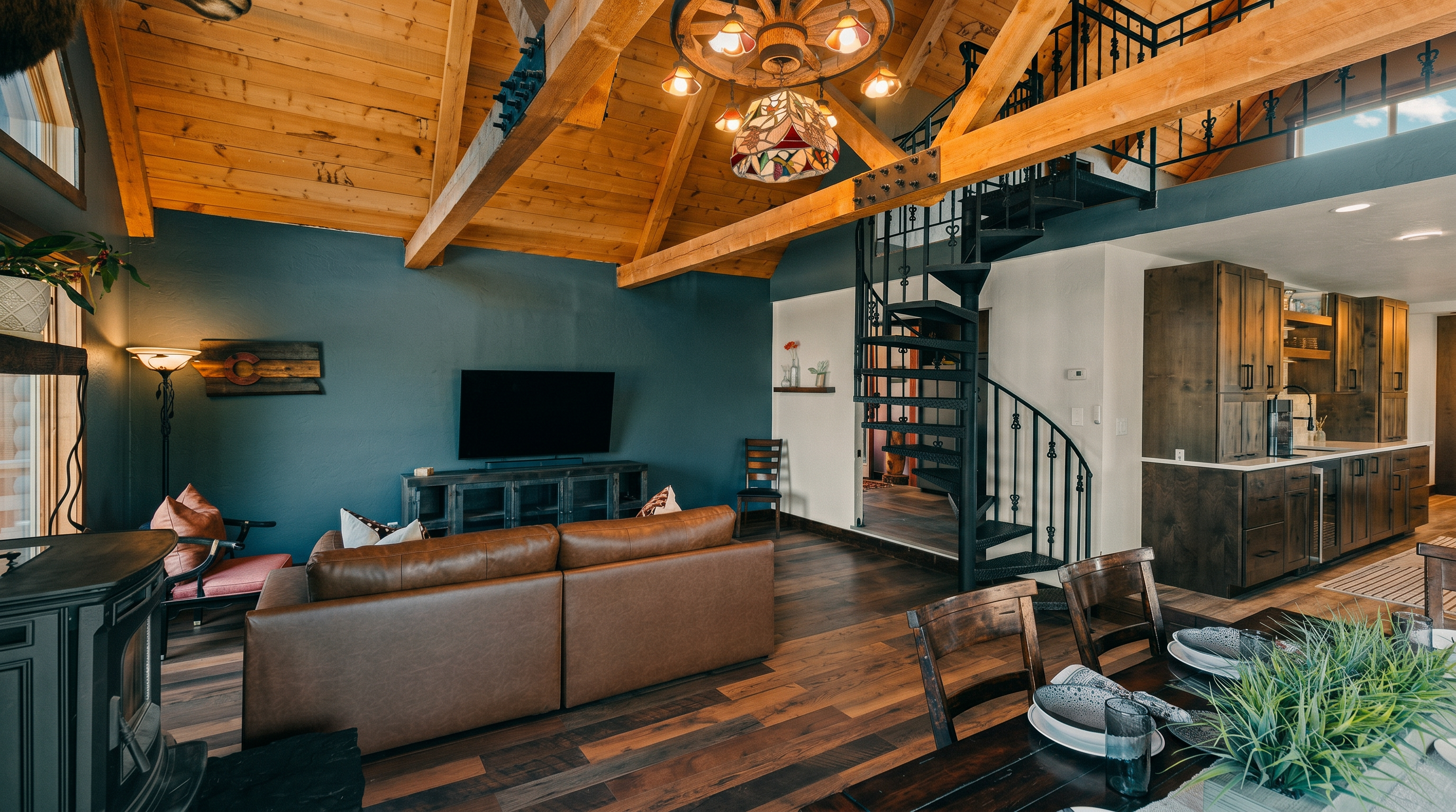 This open-concept living space features a rustic aesthetic with exposed timber beams, a vaulted wood-paneled ceiling, and a striking wagon-wheel chandelier. A leather sofa faces a wall-mounted television, while a spiral staircase leads to an upper loft area, creating a cozy, cabin-like atmosphere. The room flows seamlessly into a dining area and a kitchen with dark wood cabinetry, highlighting a warm and inviting mountain-home design.
