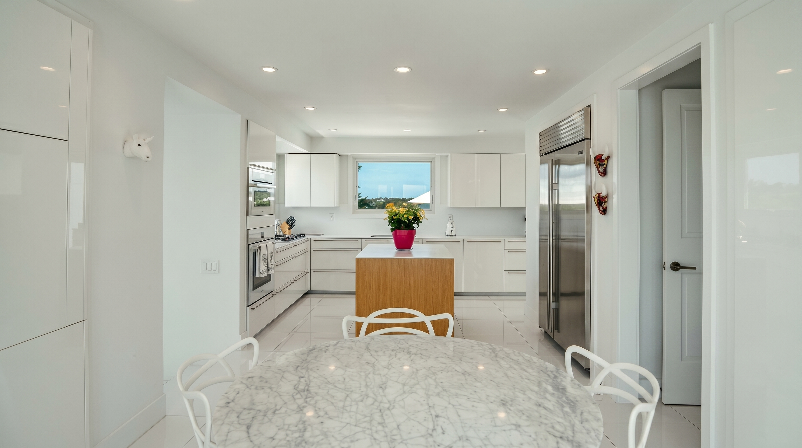 This bright, modern kitchen features a minimalist aesthetic with high-gloss white cabinetry and a central island with a light wood base. A round stone-topped dining table sits in the foreground, while a large stainless steel refrigerator and integrated appliances complete the sleek design. The space is illuminated by recessed lighting and a window above the sink that provides a view of the outdoors.