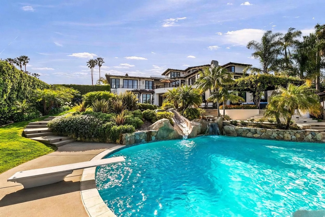 This expansive backyard features a large, curved swimming pool with a built-in rock slide and a cascading waterfall feature. A diving board extends over the water, while the surrounding landscape includes lush greenery, tiered stone stairs, and a multi-level Mediterranean-style residence in the background. The scene captures a luxurious outdoor living space designed for recreation and relaxation under a bright, clear sky.