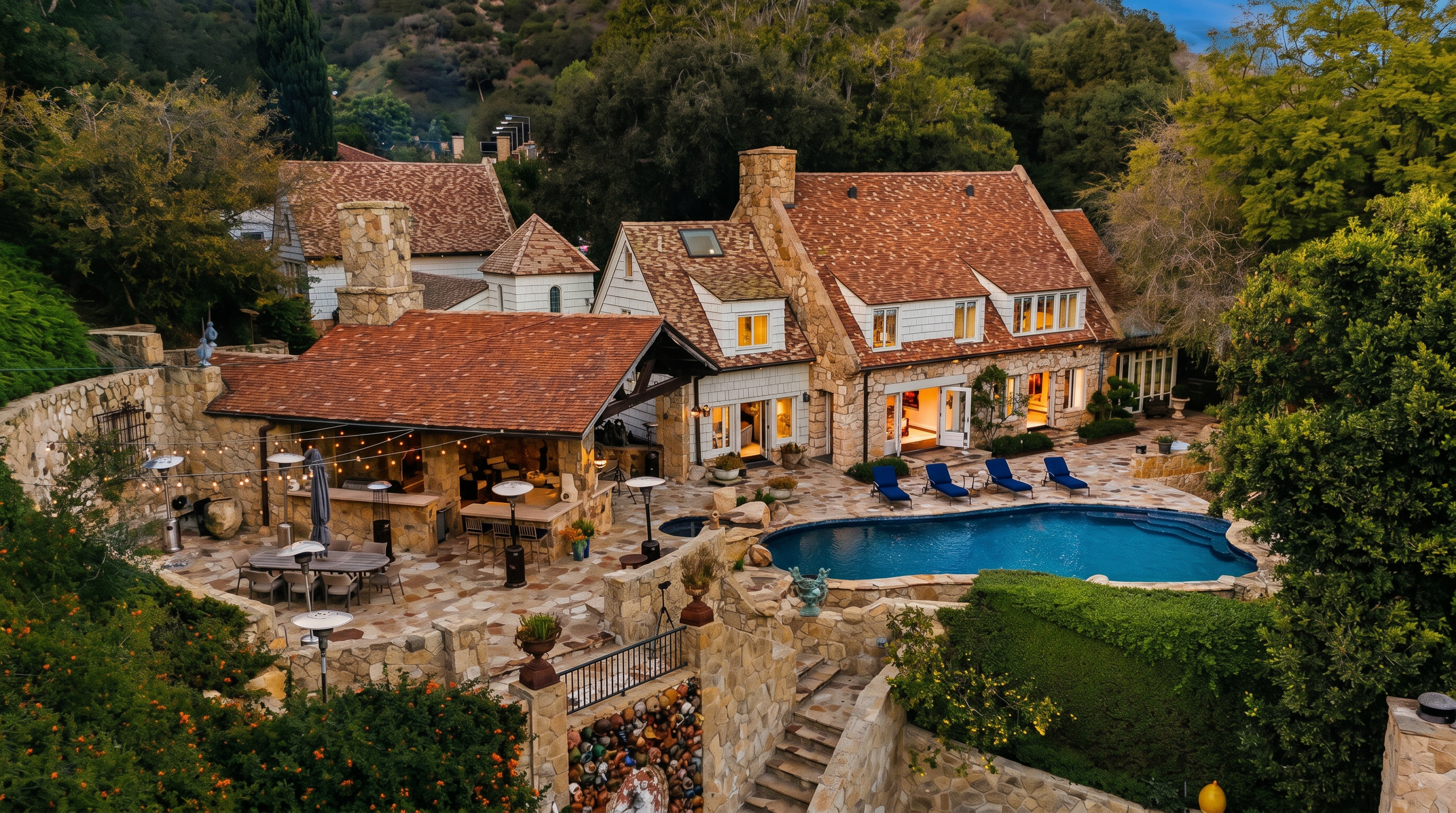 This stunning aerial perspective captures the rear of a rustic, European-inspired stone estate nestled against a lush, wooded hillside. The property features an expansive flagstone patio, a covered outdoor kitchen and dining area, and a curved swimming pool, all surrounded by mature landscaping and stone retaining walls. The home's charming architecture, characterized by its steep, multi-gabled roof and warm stone facade, creates an inviting and luxurious atmosphere.