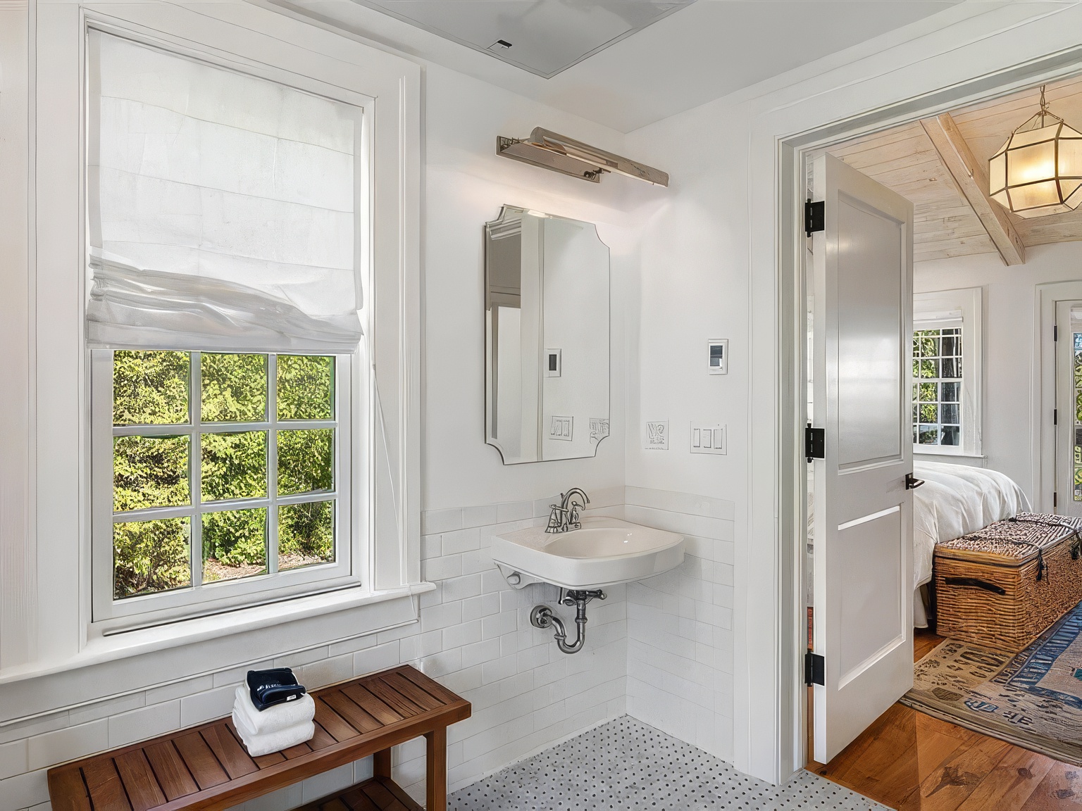 This bright, clean bathroom features a wall-mounted sink with exposed plumbing and a simple, framed mirror above. A wooden bench with stacked white towels sits beneath a window with a white roman shade, while white subway tile wainscoting lines the walls. The perspective looks from the bathroom into an adjacent bedroom, showcasing a cohesive, light-filled design with a view of a woven storage trunk and a patterned rug.