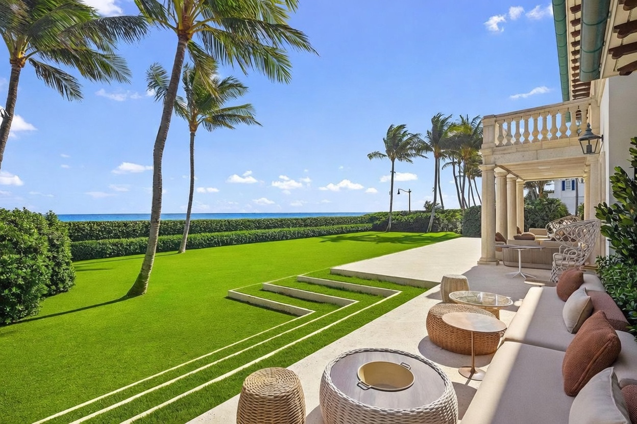 This luxurious outdoor living space features a spacious stone patio furnished with comfortable lounge seating, woven ottomans, and circular tables. The patio overlooks a meticulously manicured lawn that leads directly to a private hedge-lined boundary with an expansive ocean view. The scene is framed by tall palm trees under a clear blue sky, creating a serene and high-end coastal atmosphere.