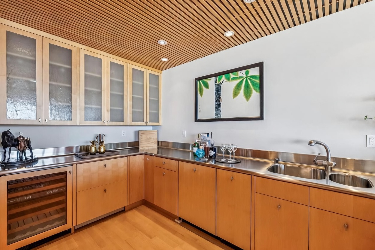 This kitchen features light wood cabinetry paired with a sleek stainless steel countertop and backsplash, creating a modern and functional workspace. The room is accented by a unique slatted wood ceiling and a framed botanical print on the wall, while a built-in wine refrigerator and a double-basin sink add convenience. The perspective is a wide-angle shot capturing the L-shaped layout, emphasizing the clean lines and contemporary aesthetic of the space.