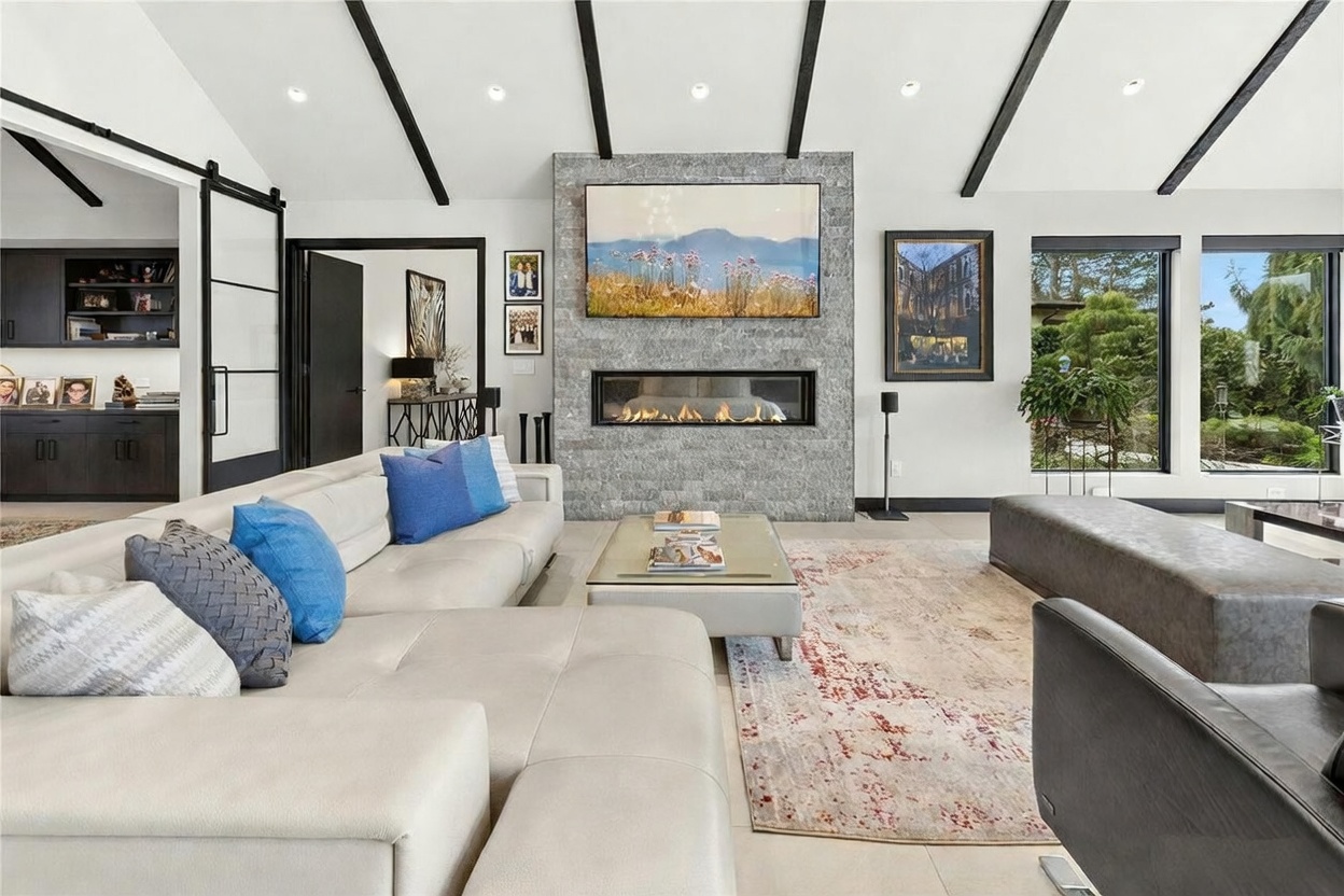 This spacious, modern living room features a striking floor-to-ceiling stone fireplace with a built-in linear gas insert and a large television mounted above. The room is anchored by a large, light-colored sectional sofa and a patterned area rug, set against a backdrop of vaulted ceilings with dark exposed beams. Large windows provide ample natural light and a view of the lush greenery outside, creating a sophisticated and inviting atmosphere.