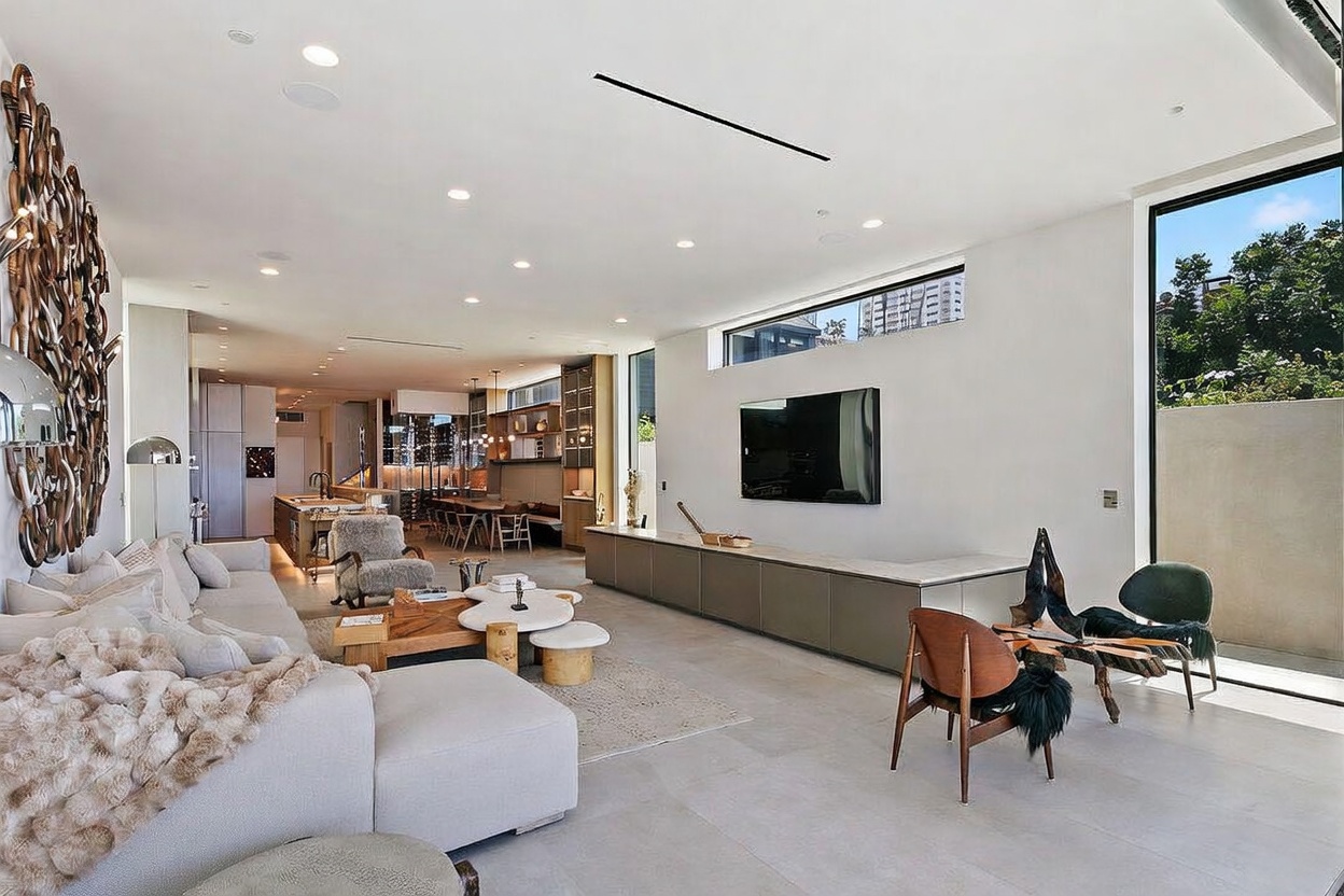 This expansive, open-concept living room features a minimalist aesthetic with neutral tones and large-format tile flooring. The space is anchored by a low-profile media console beneath a wall-mounted television, complemented by a textured area rug and a unique, multi-tiered coffee table. Large windows and a glass door provide abundant natural light, while a striking, intricate wall sculpture adds a dramatic artistic focal point to the room.